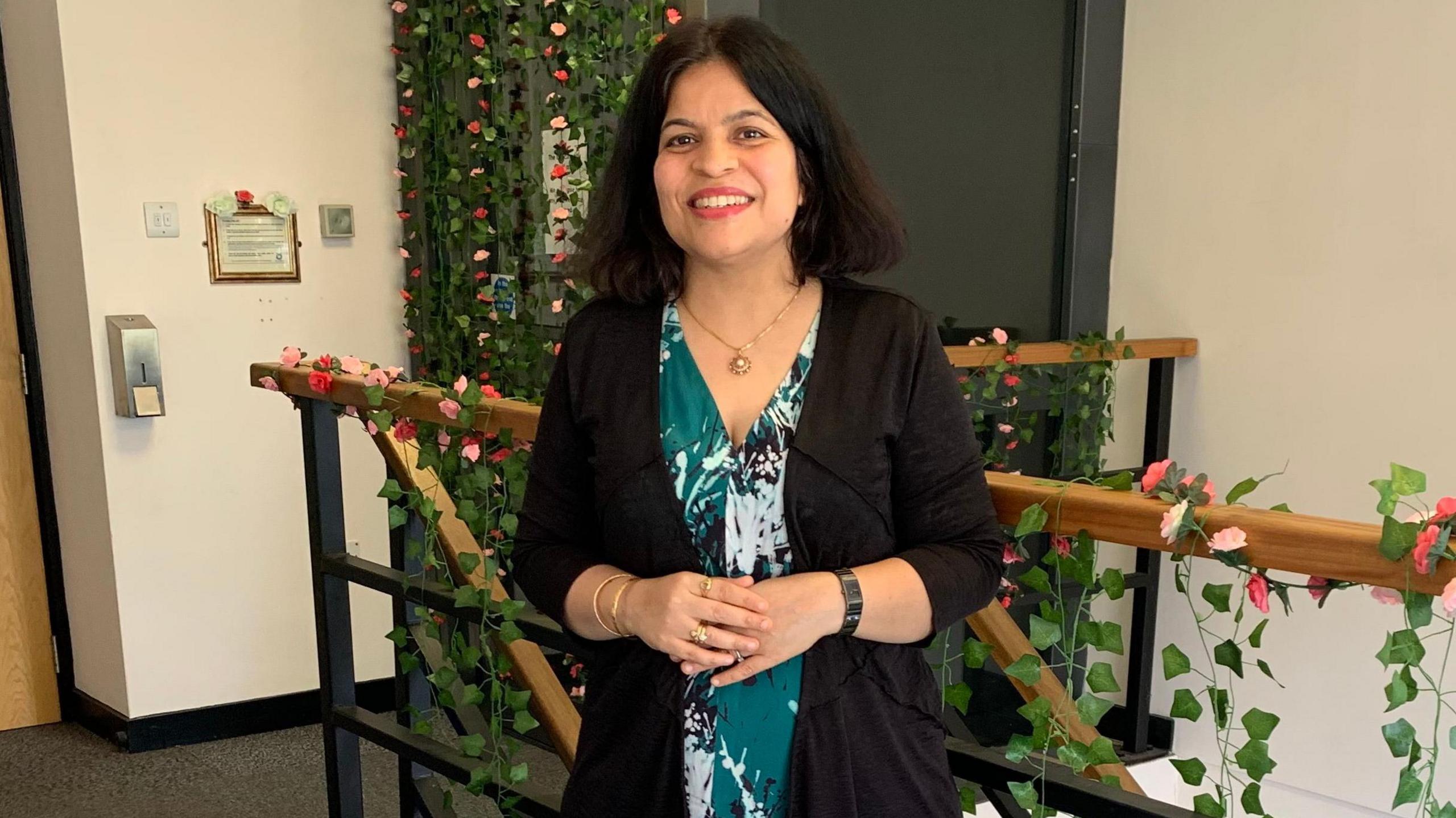 An Indian woman stands at the top of a staircase which is decorated with trailing green leaves and pink flowers. She is wearing a dark green dress with a white pattern and a black cardigan. she has dark hair in a long bob and is smiling into the camera.