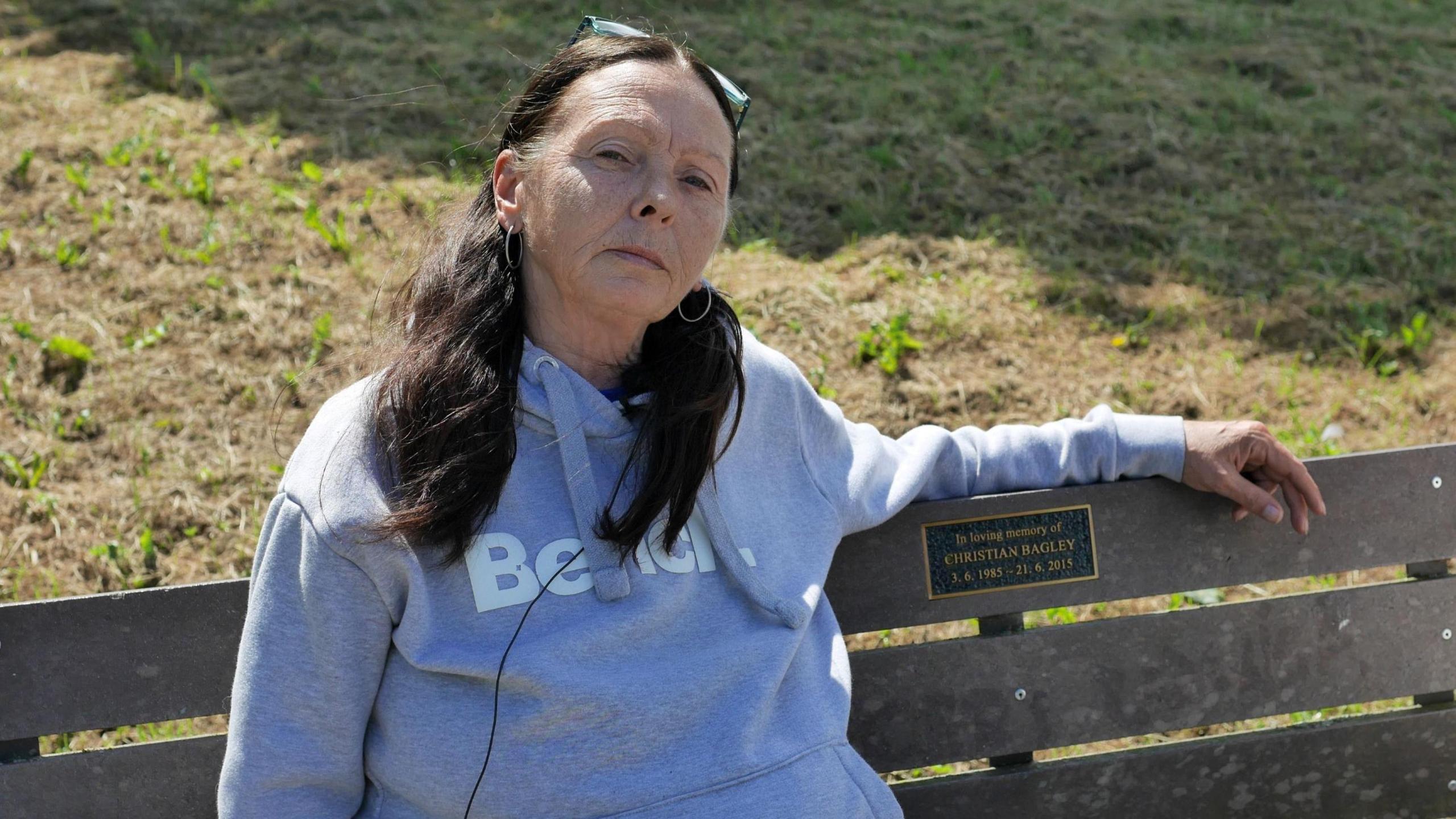 A woman with long dark hair in pigtails, wearing a grey Bench hoodie, sits on a wooden bench. A plaque on the bench reads: "In loving memory of Christian Bagley - 3.6.1985-21.6.2015".