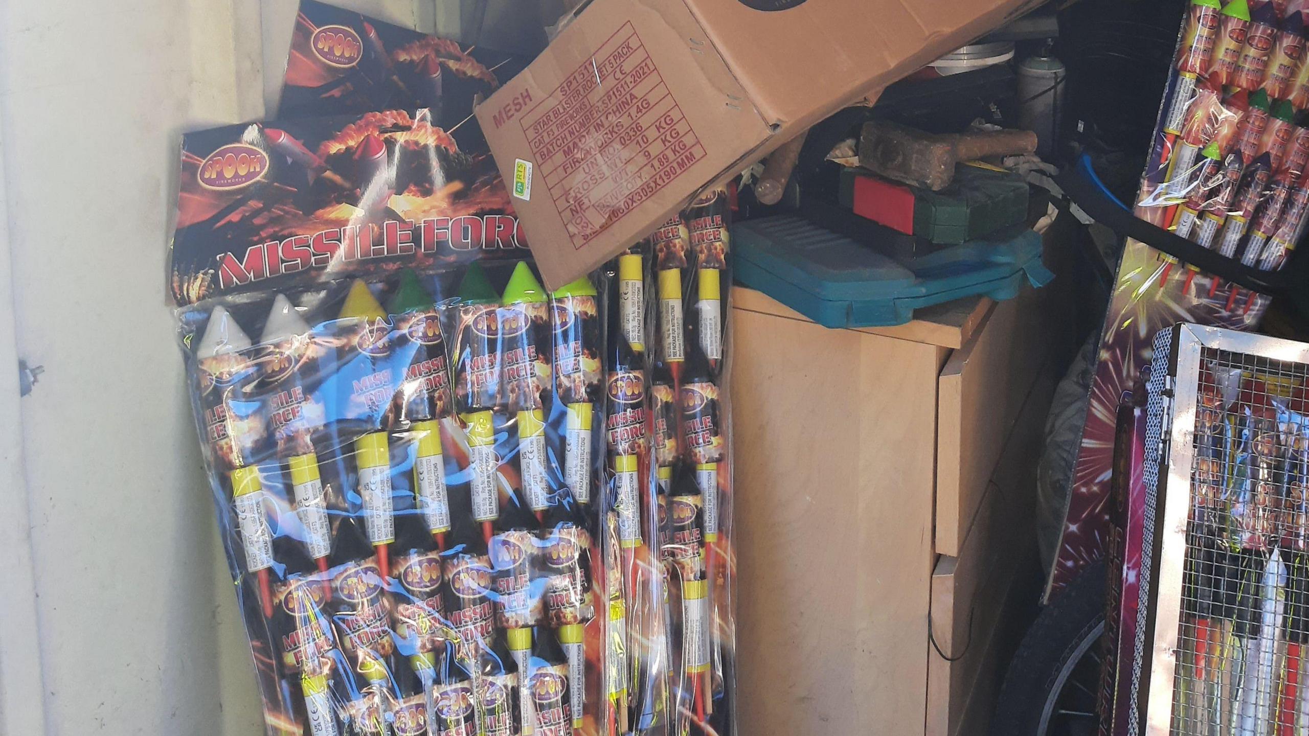 Packets of rocket fireworks, with the words 'missile force' written on the label and on each firework, are propped up against a chest of drawers. A cardboard box is lying over the top of the packets. More packets of rocket fireworks are in the background.