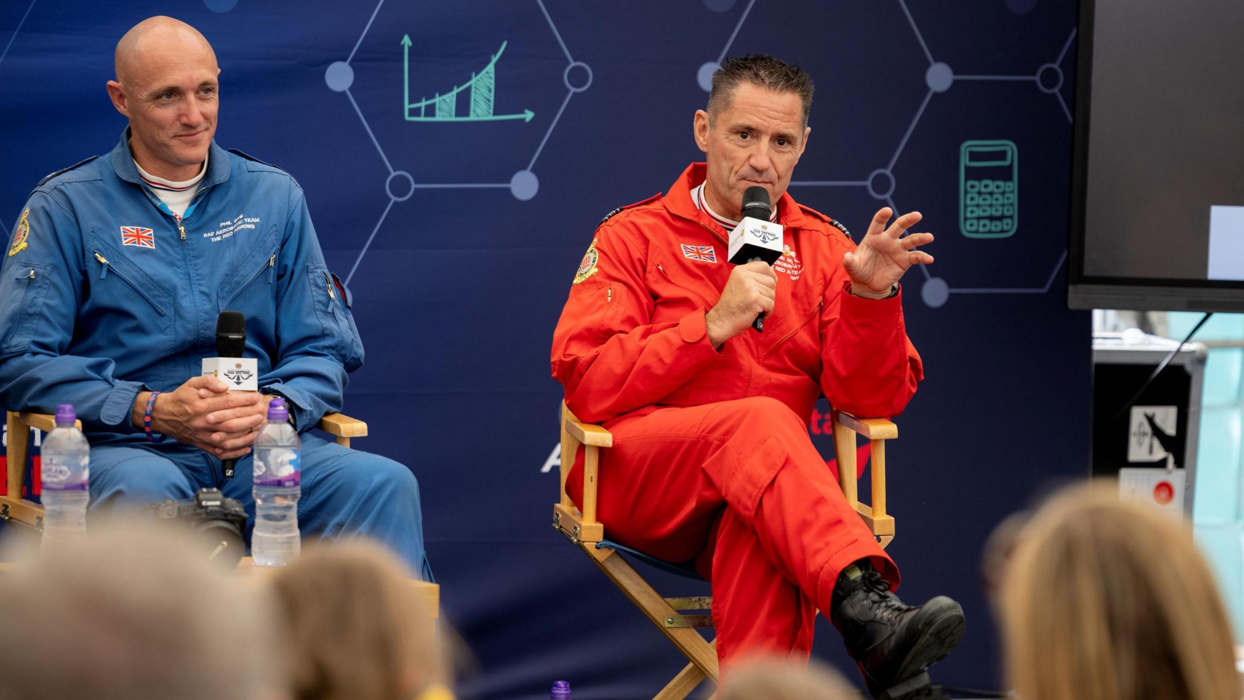 A man in a red flight suit sitting on a chair speaking into a microphone, sitting next to a man in a blue flight suit