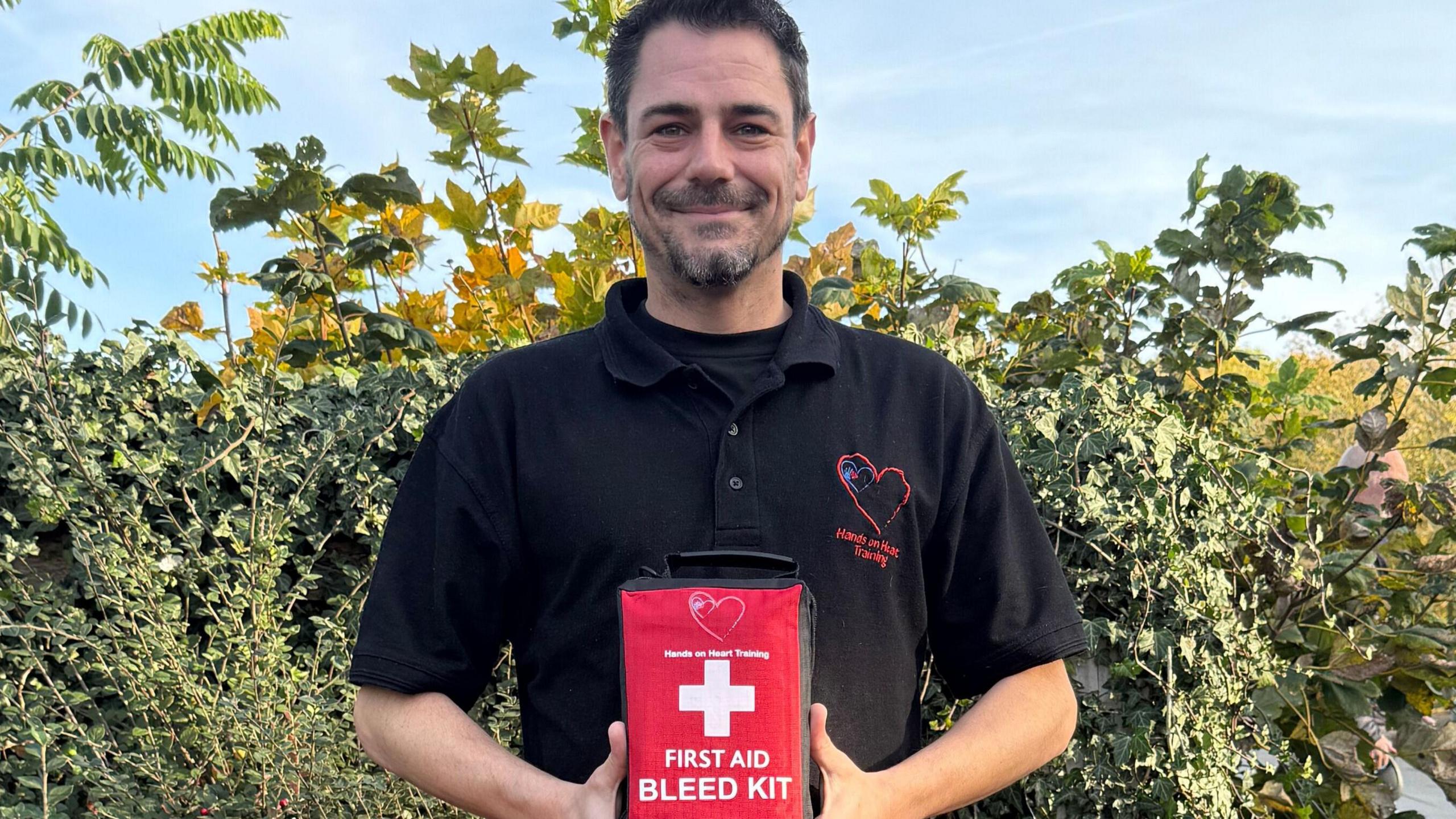 A man is holding a red first aid bleed kit. He is wearing a black polo with the logo "Hands on Heart Training". Behind him are green bushes and a clear, blue sky.