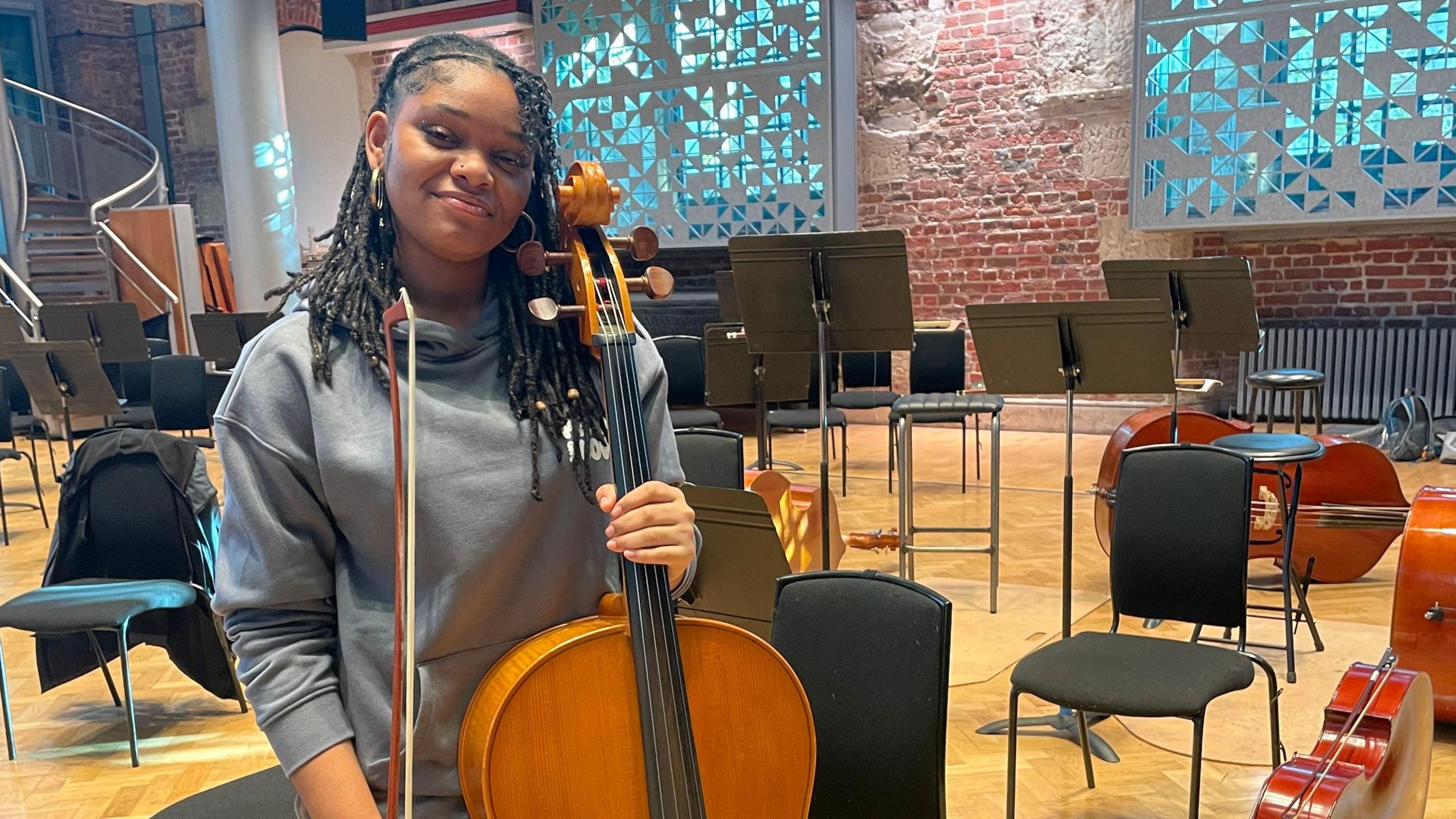 Ayaana is standing up with the cello in her hand. She is smiling with a grey hoodie on. In the background there are scattered chairs and instruments. 