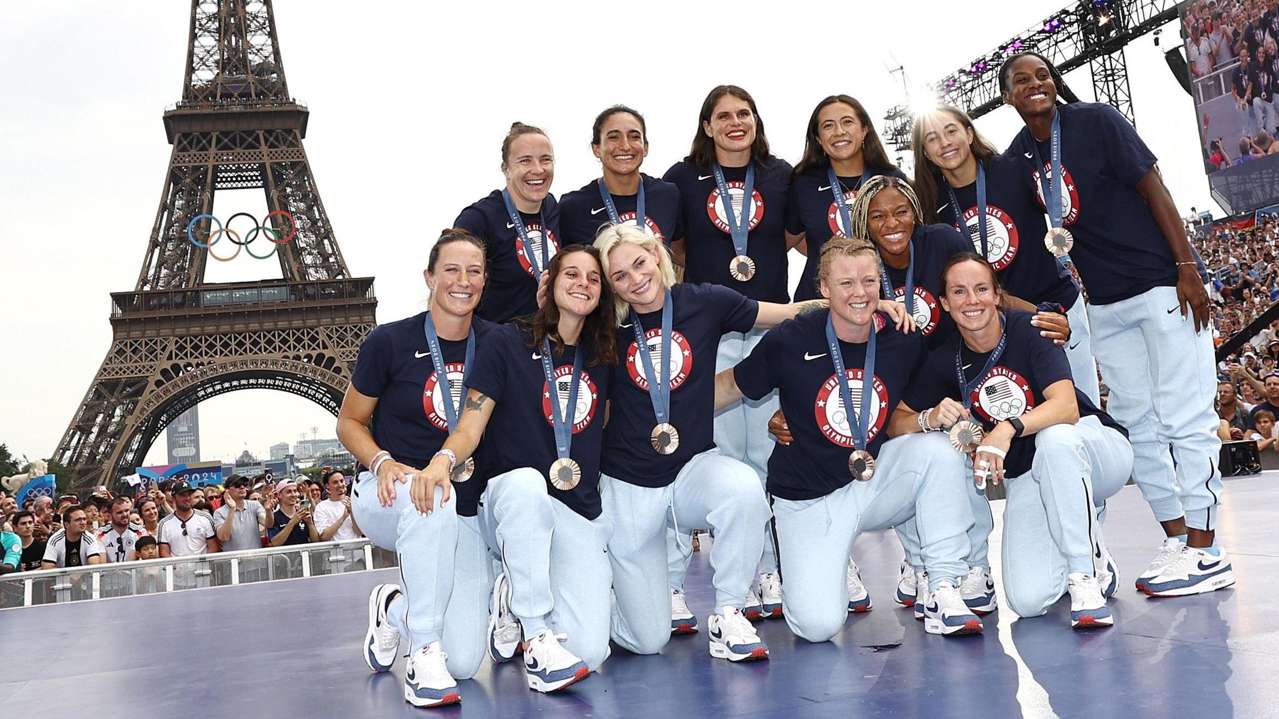 12 members of the USA women's rugby sevens team gather together to pose with bronze medals around their necks at the Paris 2024 Olympics. They are on a blue raised platform which has a crowd around it, while in the background can be seen the lower half of the Eiffel Tower with the famous Olympic rings logo suspended from it