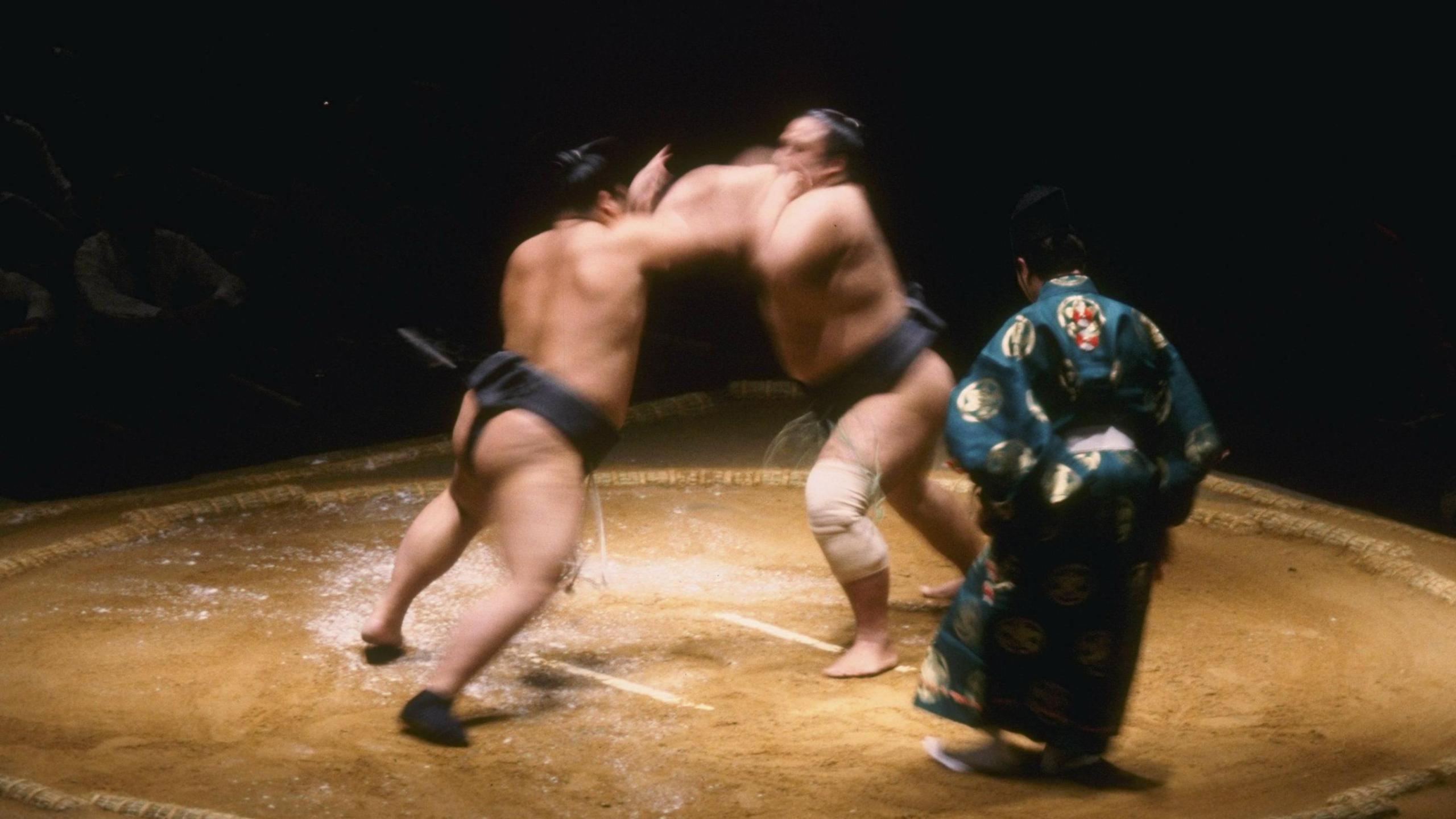 Two sumo wrestlers fighting each other in a ring with a referee in traditional dress mediating the match.