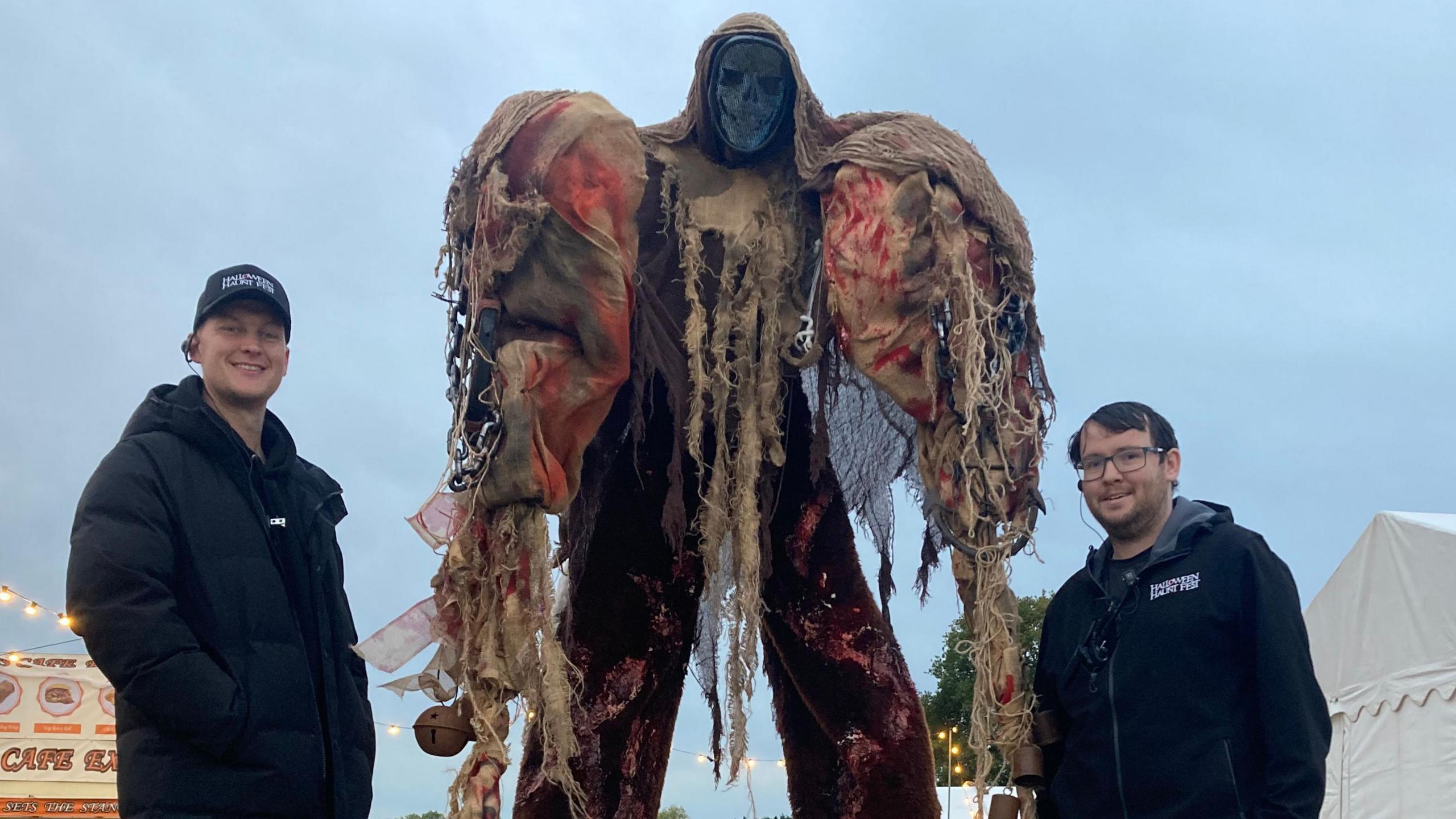 Two men in branded Halloween Haunt Fest hats and coats stand either side of a tall performer dressed as a grey faced skeleton
