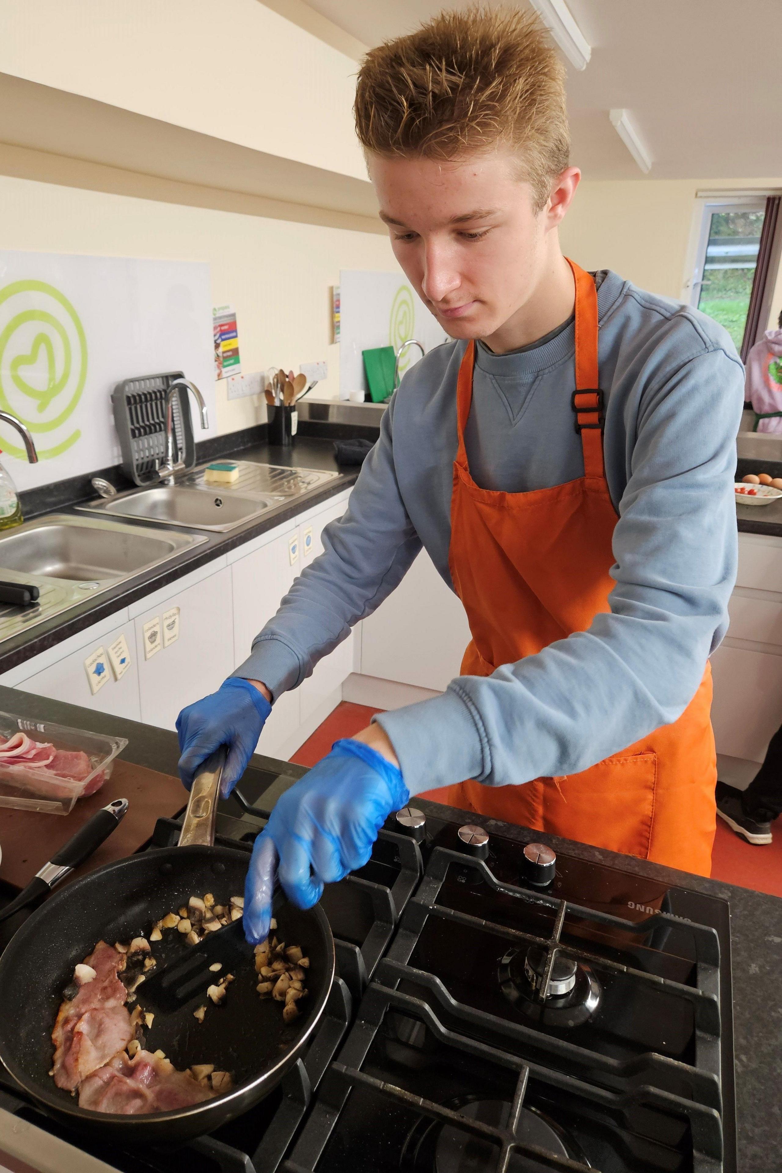 James, wearing an orange apron frying bacon on a hob in a kitchen