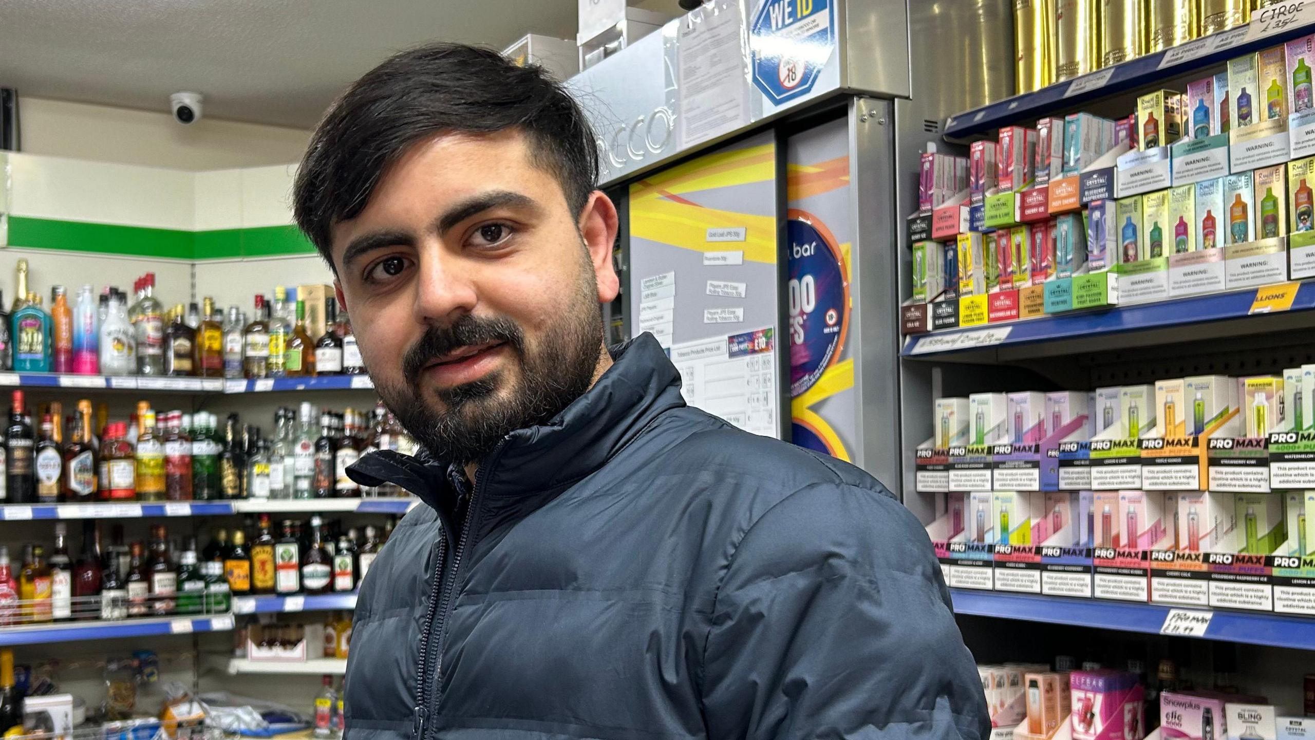 A man wearing a blue coat which is zipped up. He has a beard and moustache. He is standing inside a shop with liquors and vaping supplies visible on the shelf behind him