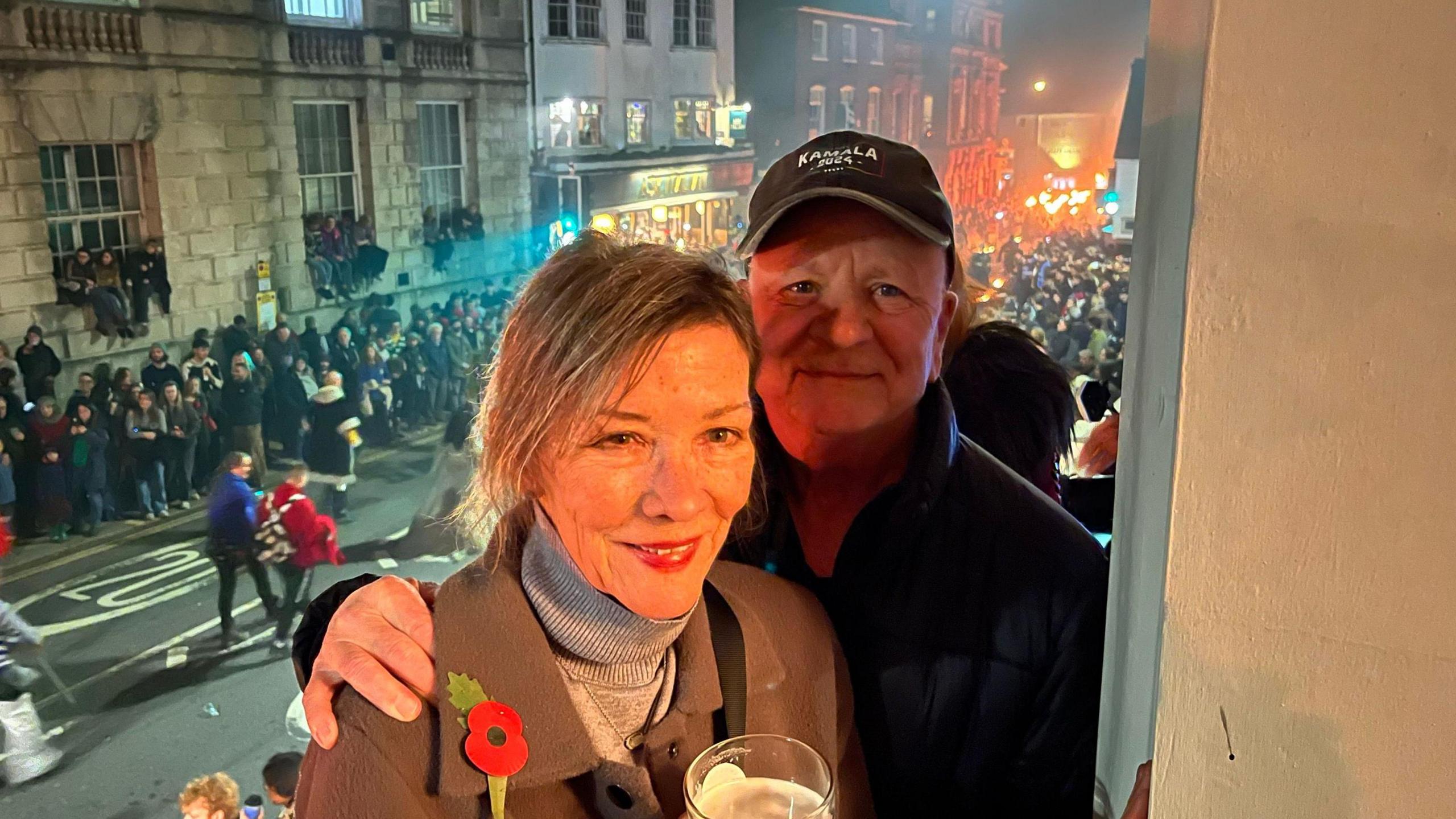 A man and a woman facing the camera. Behind them are the Lewes Bonfire celebrations on the street below