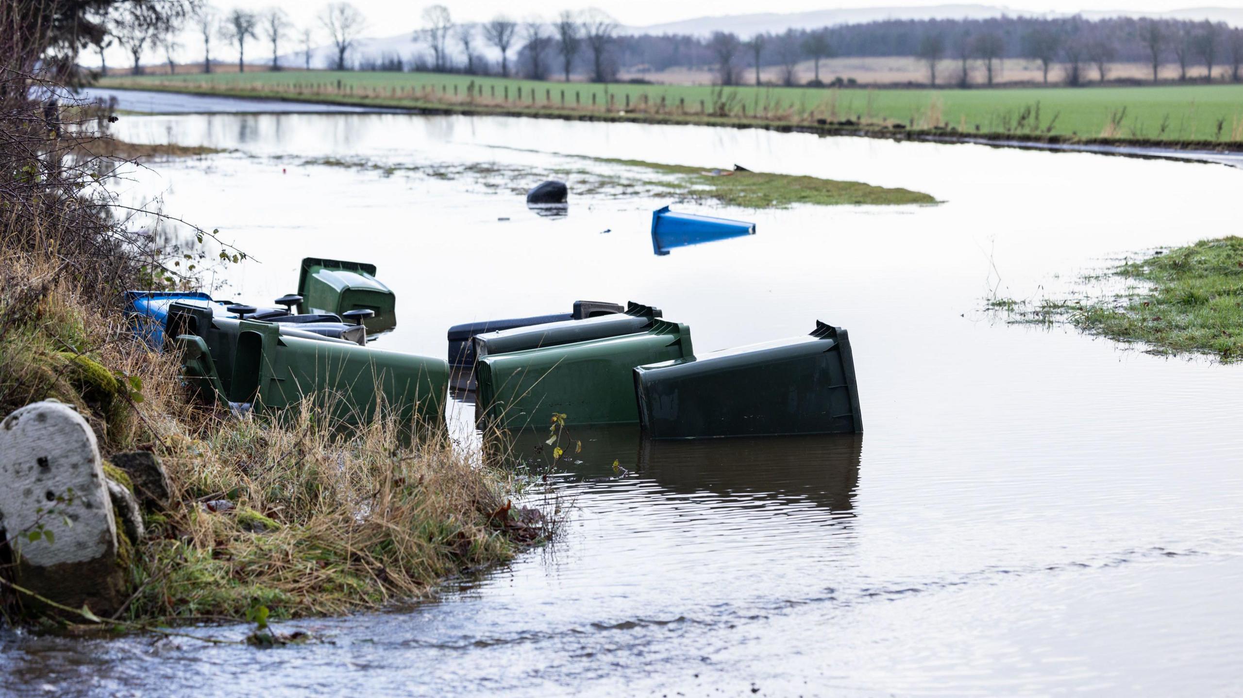 Several partially submerged wheelie bins were toppled over by flooding on the A9 near Invergordon. The road is under water apart from small patches of grass and fields can be seen in the background.