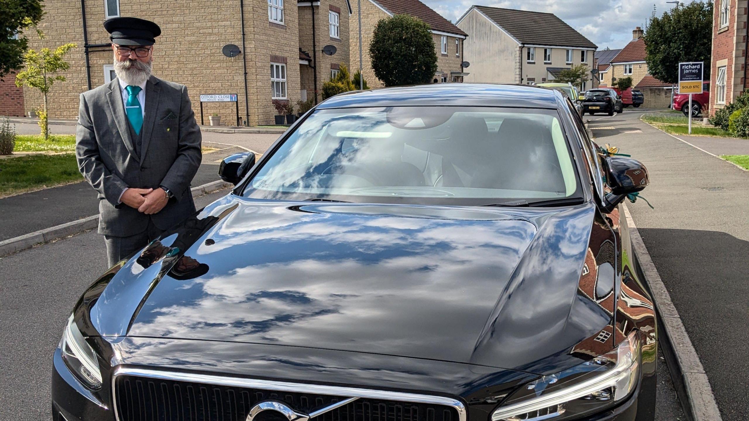 The front of a black, shiny limo parked in a residential area. To the side, the driver stands next to it smiling at the camera. He is wearing a chauffeur-type outfit, complete with hat and a blue tie.