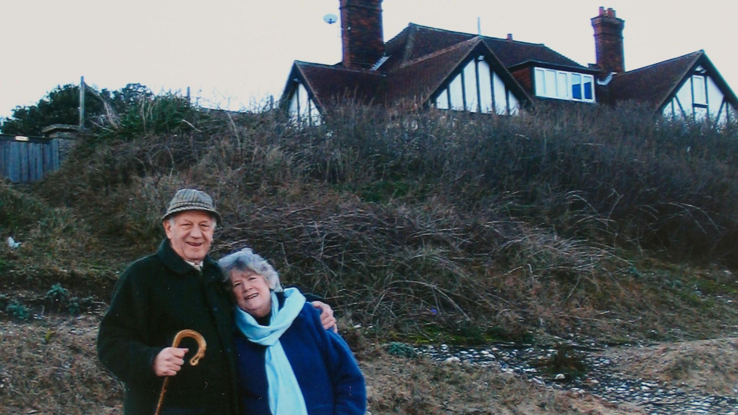 Jean Flick with her husband at their home in 2013. They are smiling at the camera and standing arm in arm. They wear winter coats and her husband wears a hat and a walking stick.
