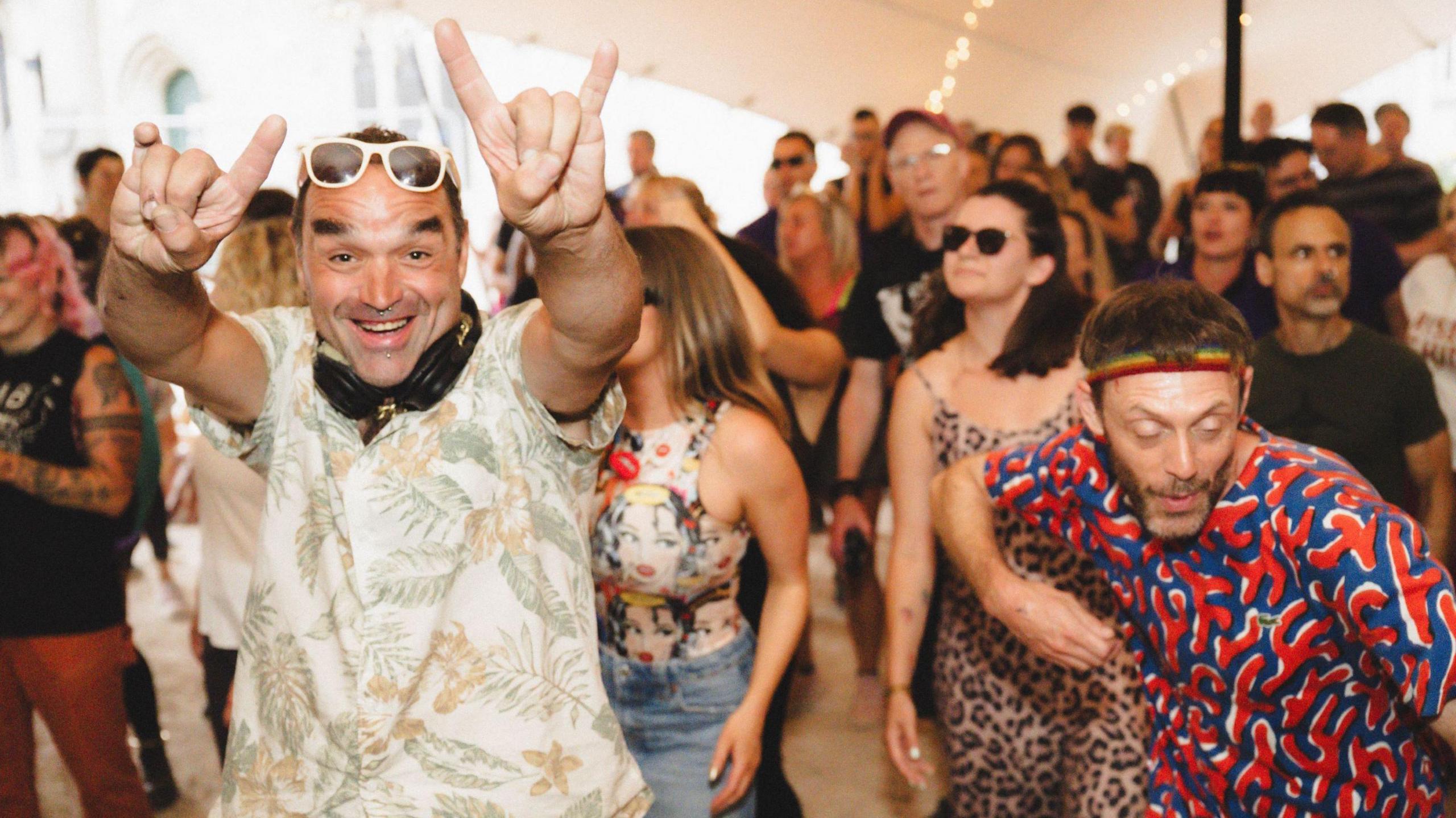 A group of people dance inside a teepee at a sober rave event in Bristol. Closest to the camera are two men, one wearing a light-coloured patterned short-sleeved shirt, he is waving at the camera and smiling and has sunglasses at the top of his head. The other man, wearing a blue and red patterned top and a multi-coloured headband, is looking down as he dances.