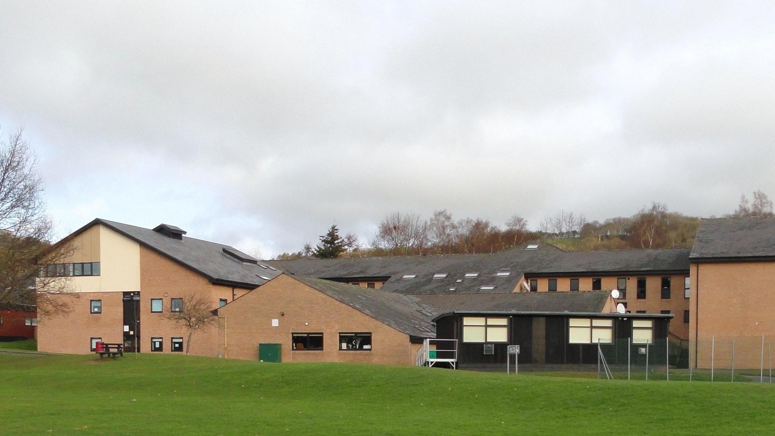 A general view of Crickhowell High School, a group of red brick buildings with a grass bank in front of it and a metal fence to the right. Trees can be seen in the horizon.