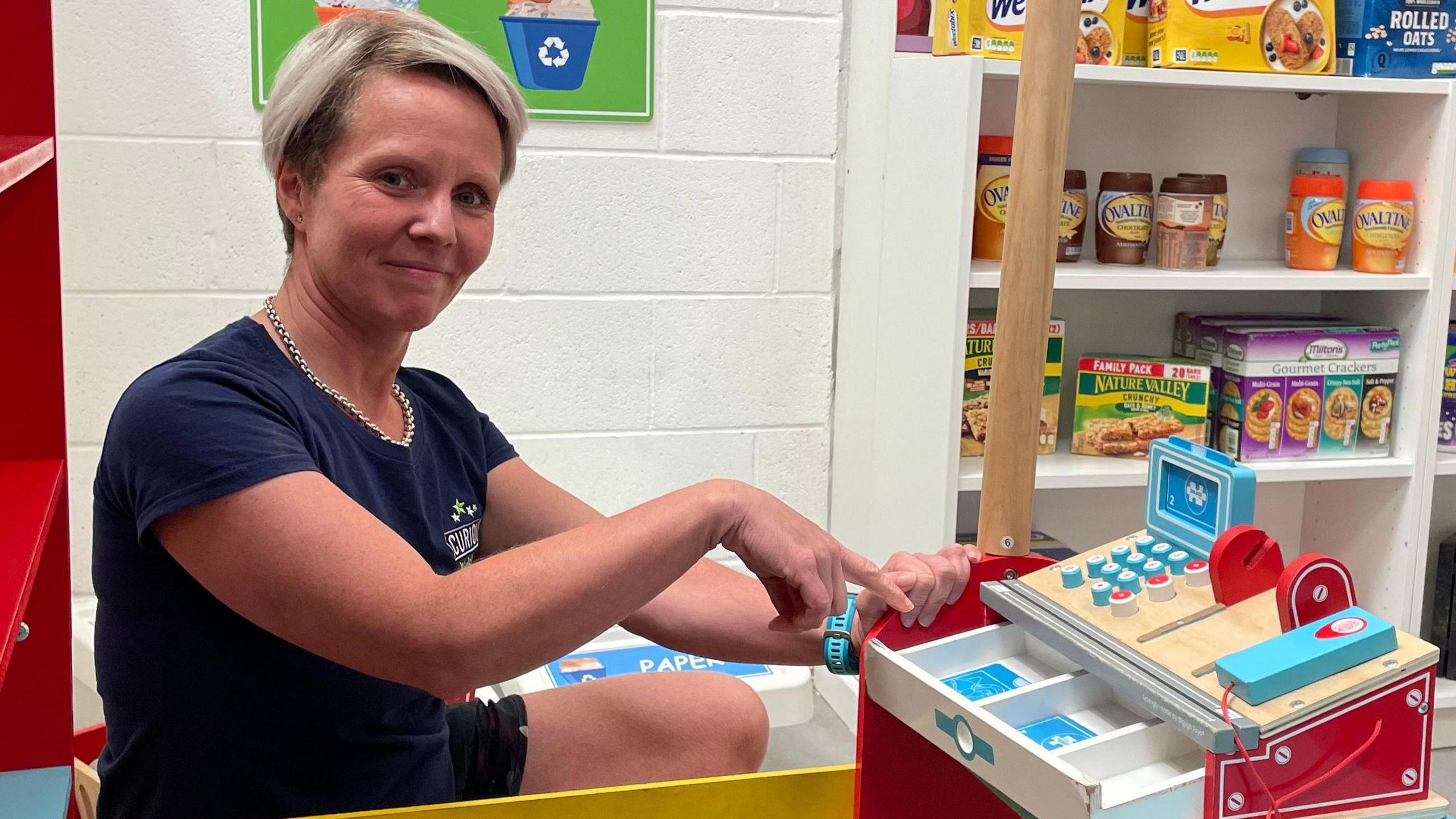 Rachael Parker pointing into an empty toy cash register at the play area, inside a mock-up supermarket designed for children's roleplay.
