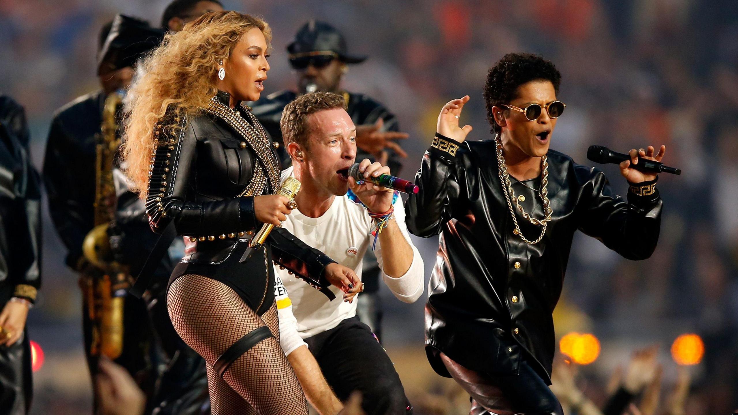 Beyonce performing at the 2016 Super Bowl half-time show alongside Coldplay's Chris Martin and Bruno Mars. Beyonce is styled in a leather body suit with fish net tights, her hair long and curly. Martin wears a white top and black jeans while Mars matches Beyonce in black leather, sunglasses and gold accessories. 