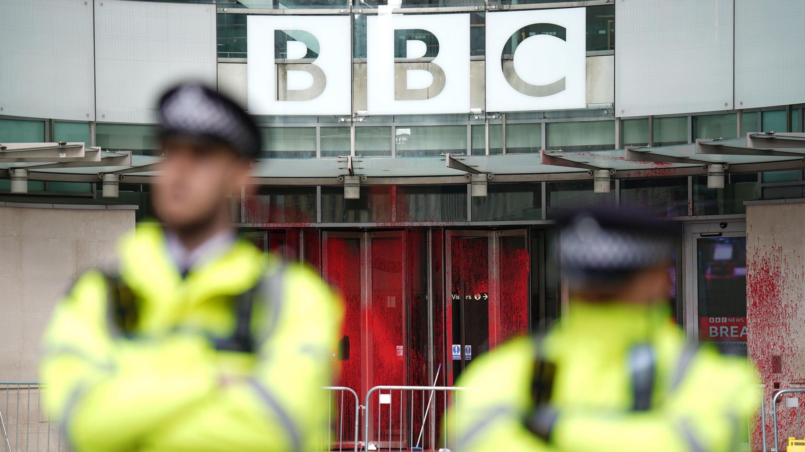 Image showing the paint splattered across the three revolving doors at the entrance to New Broadcasting House, while two out-of-focus uniformed police officers stand side by side 