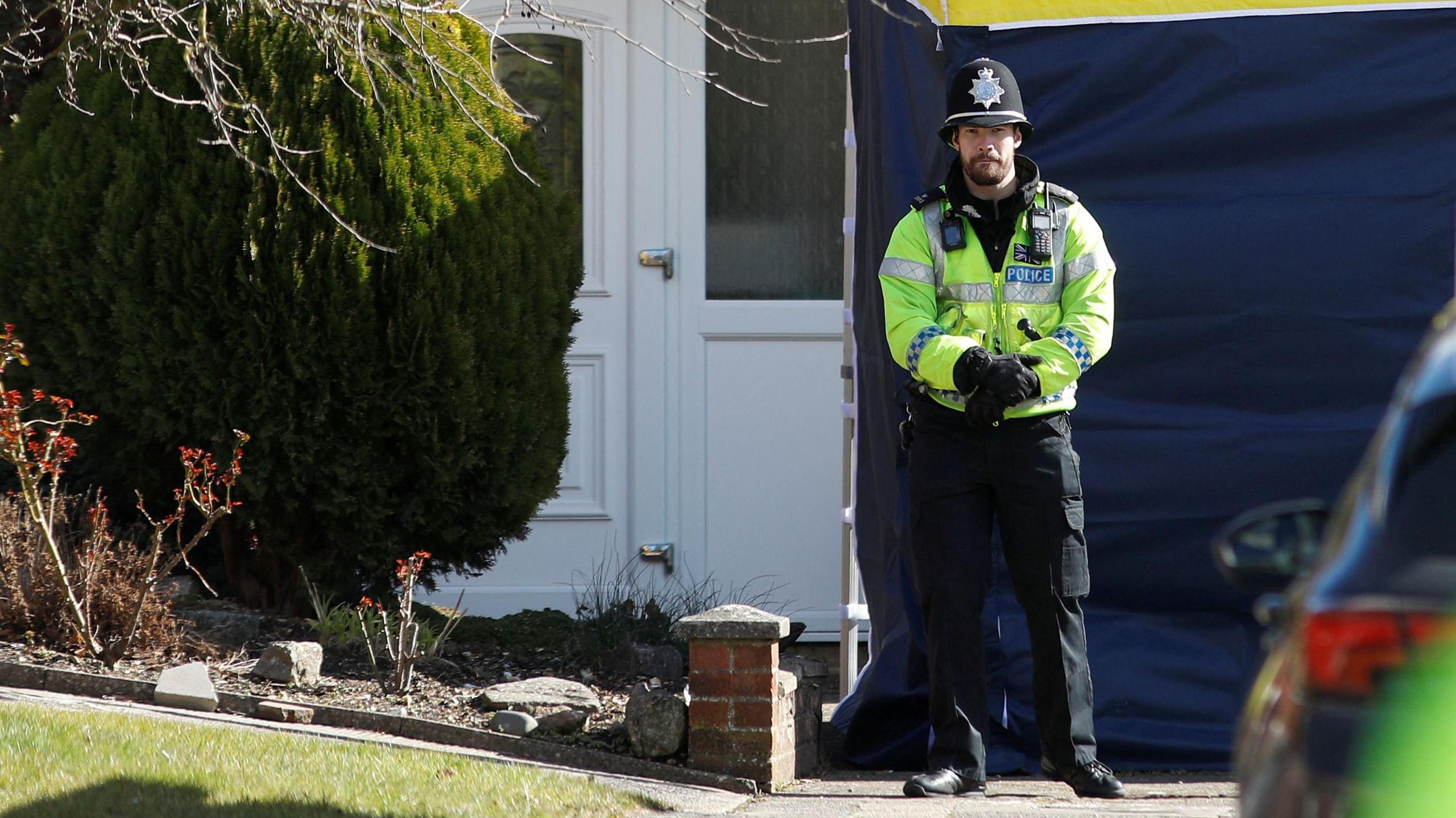 A male police officer standing outside Sergei Skripal's house in Salisbury, Wiltshire, in 2018. The officer is wearing a yellow high vis police jacket, a black hat with a police emblem, black trousers and black gloves. He has dark facial hair. A police tent can be seen behind him. 