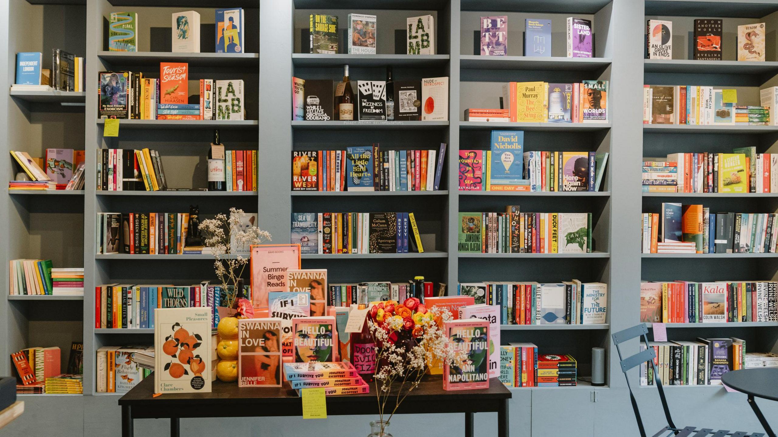 An interior view of Bard Books. Colourful books are stacked on shelves. Books and flowers also cover a table in front of the shelves.