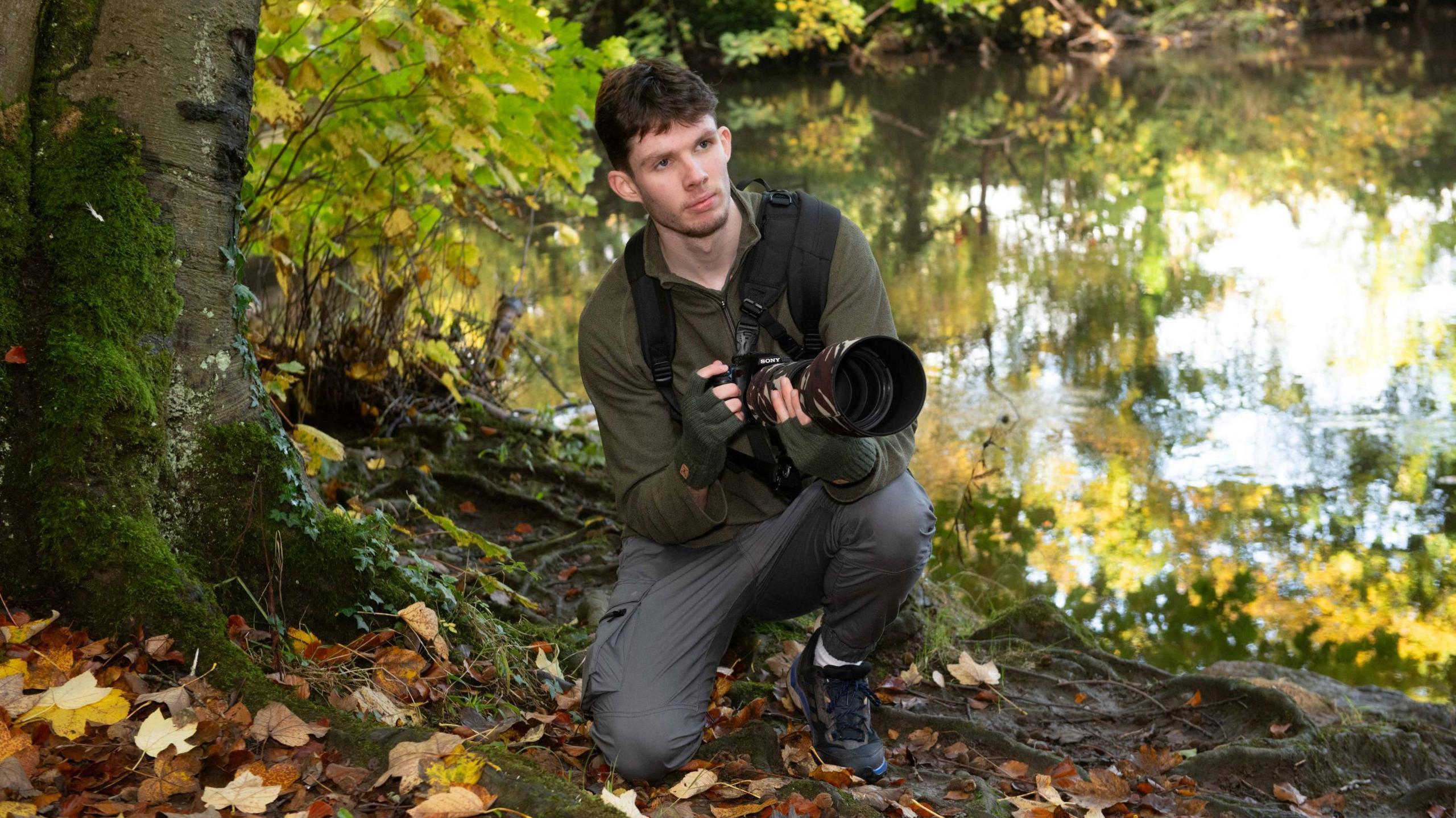 Liam Pinchen holding a camera while crouched in front of a lake.