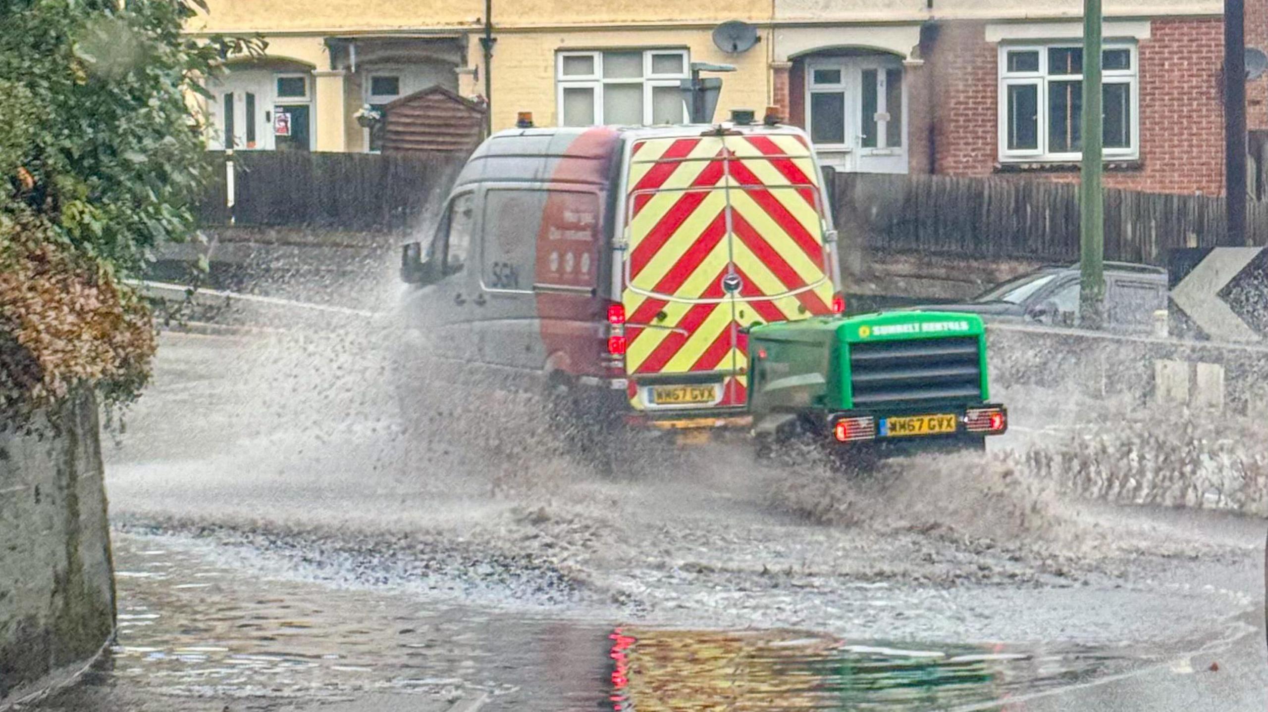 A van drives through floodwater with houses visible in the background