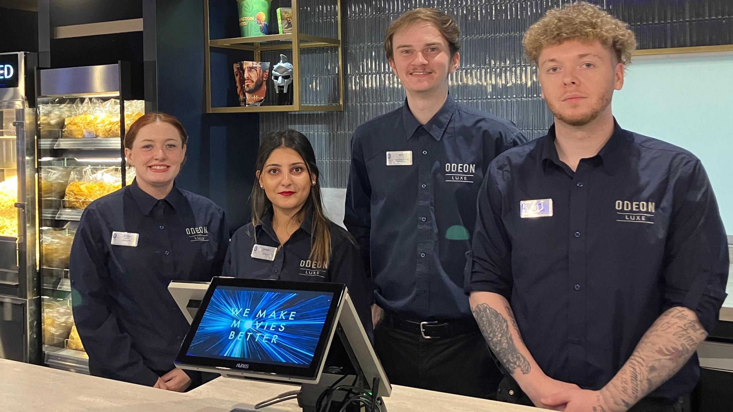 Four members of Odeon staff pose behind the bar for a picture. There are two women and two men, all wearing navy shirts. Behind them is a popcorn machine and in front of them is a cash register. 