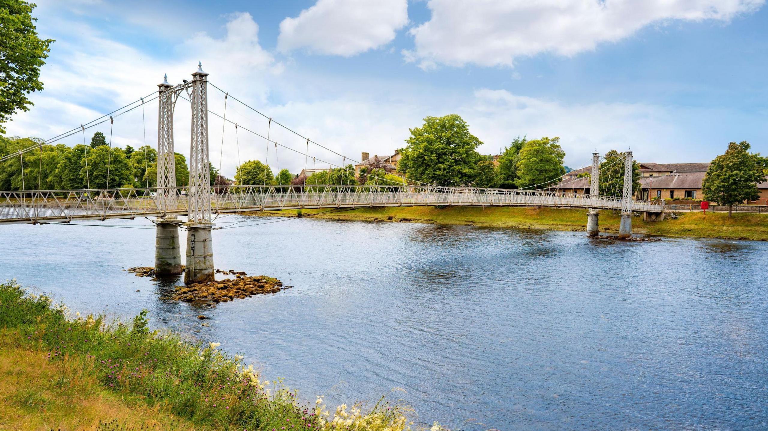 The footbridge is constructed of latticed metalwork and steel cables. The River Ness below is at a low level and brown stones are visible around the piles supporting the bridge. Trees line a bank of the river.