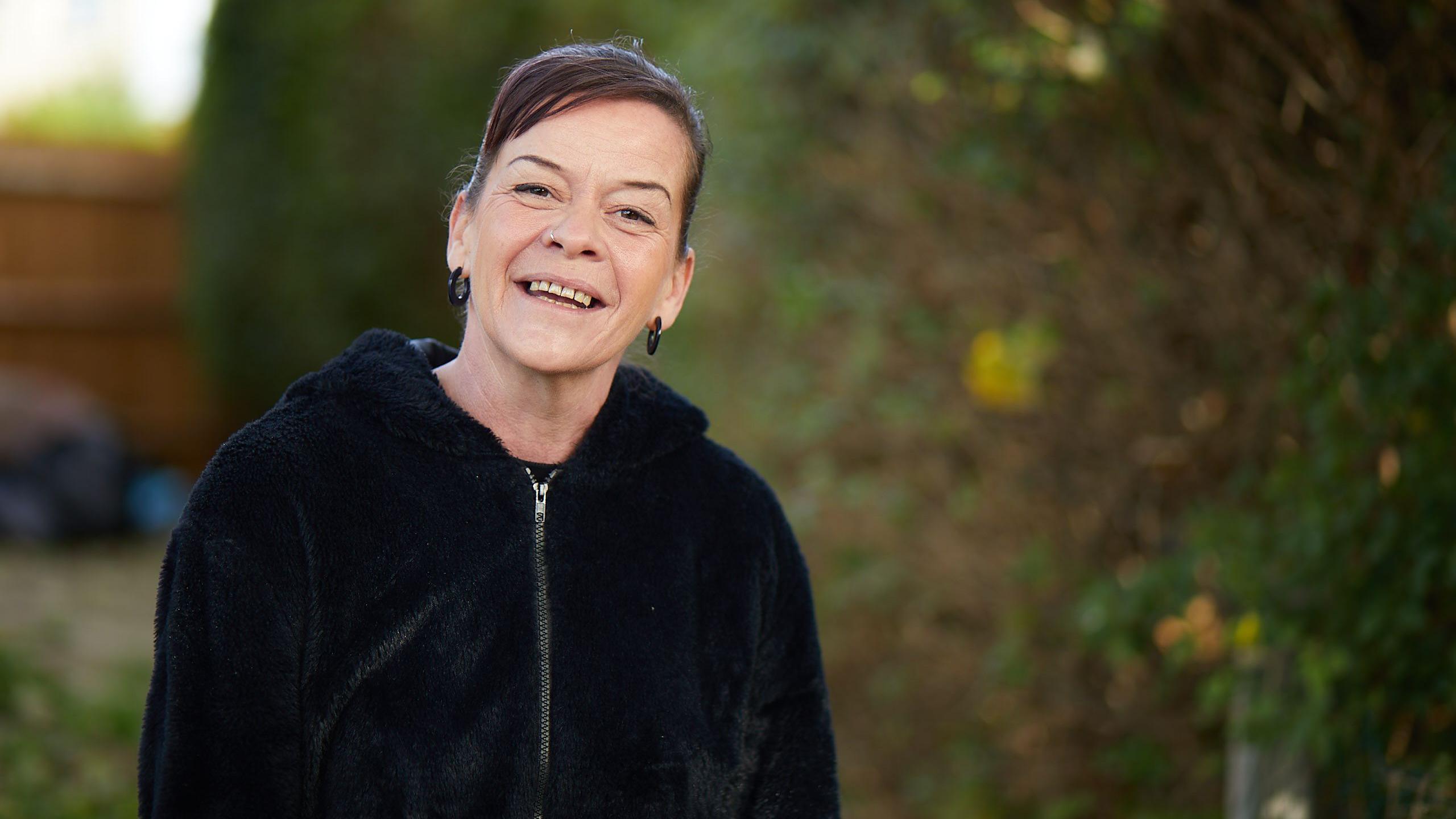 Amanda Wall. She is photographed outside, wearing a black zip jacket. She has dark hair which is tied up and is wearing black hoop earrings. She is smiling at the camera. 