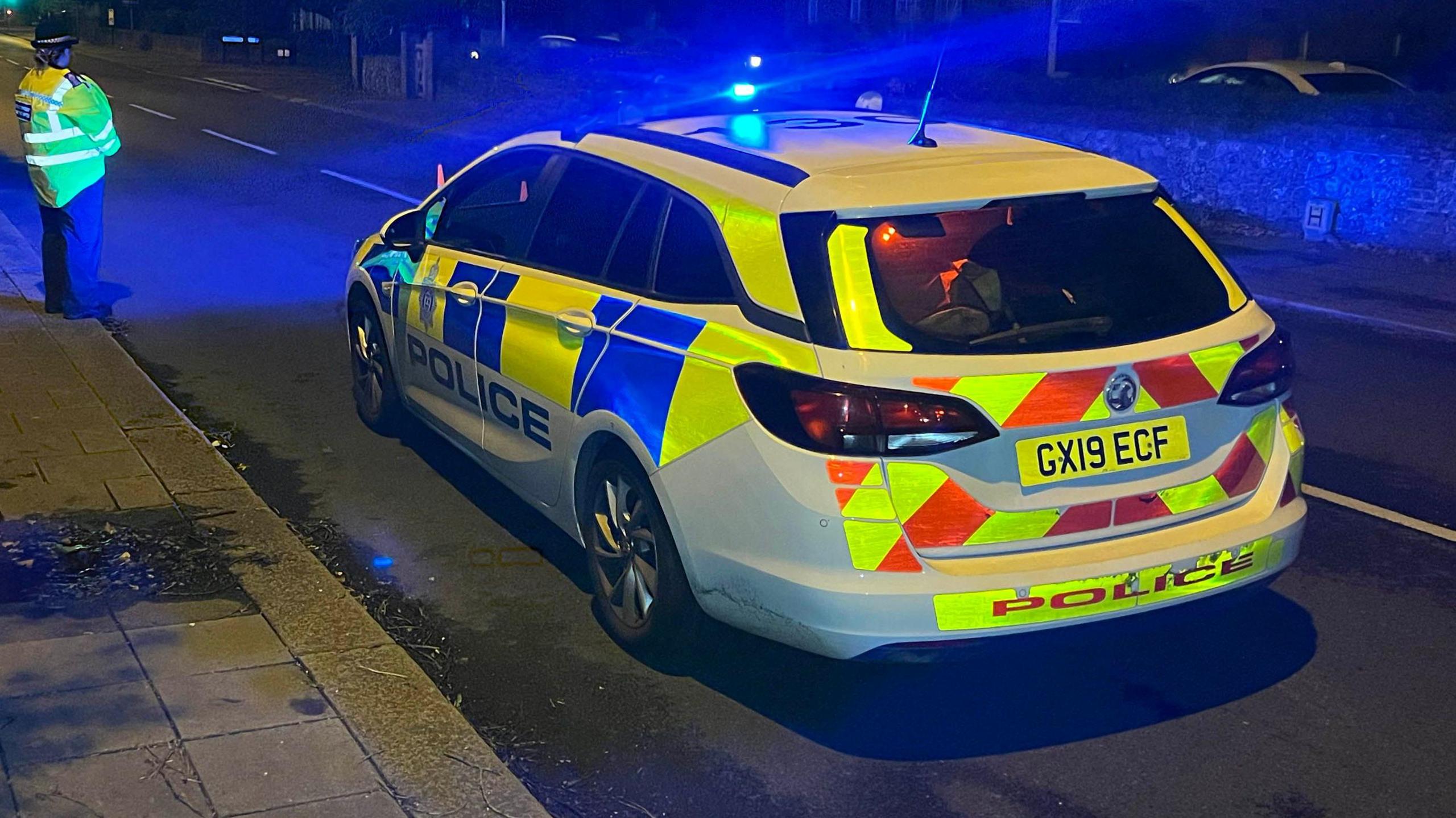 A police car is parked in a road, with a female officer standing on the pavement next to the vehicle. It is dark and the police car has blue lights. 