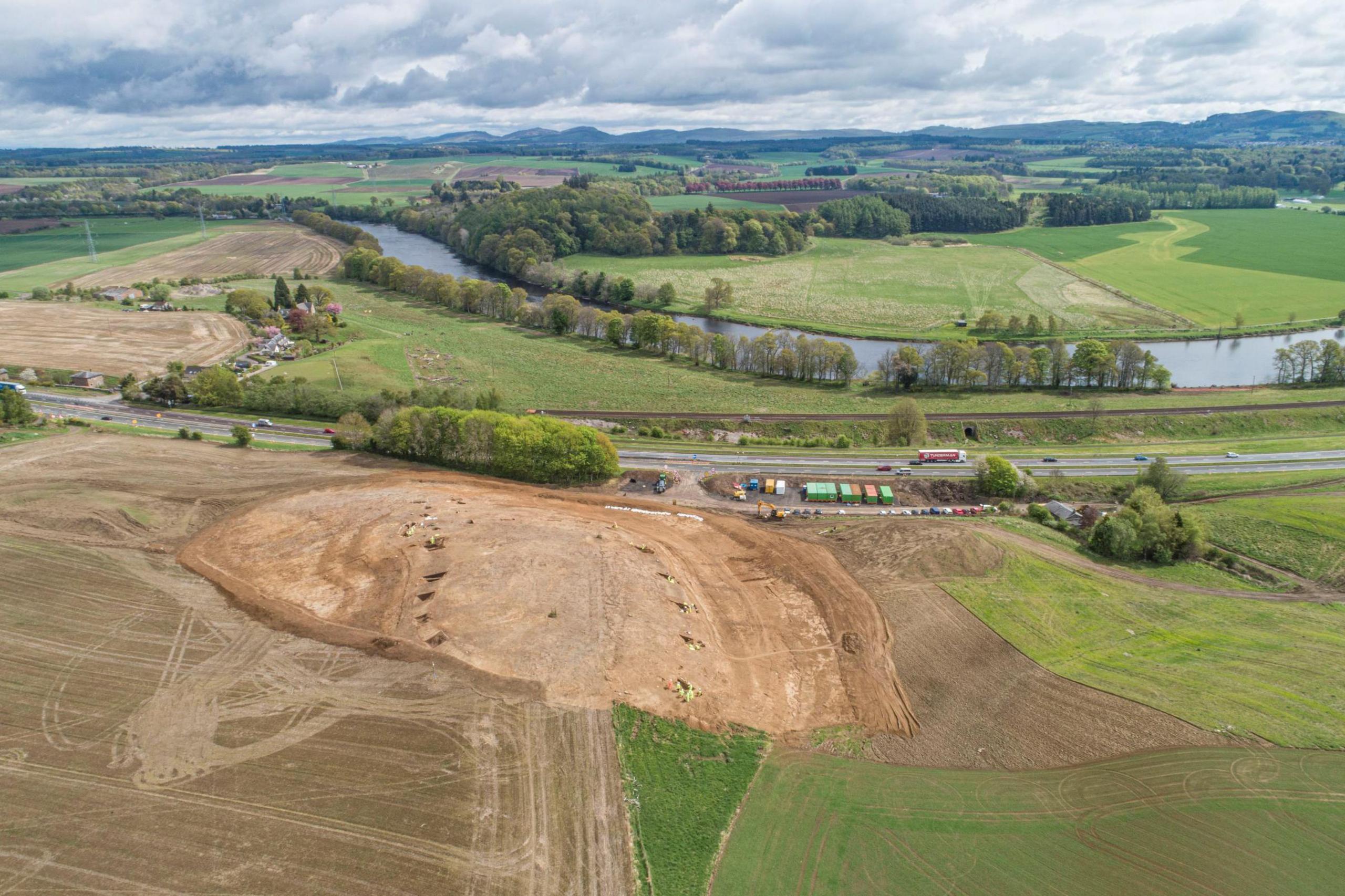 Aerial view of Broxy Kennels Fort under excavation. A large area of a field has been excavated. The site is in an area of fields and trees and the river Tay. 