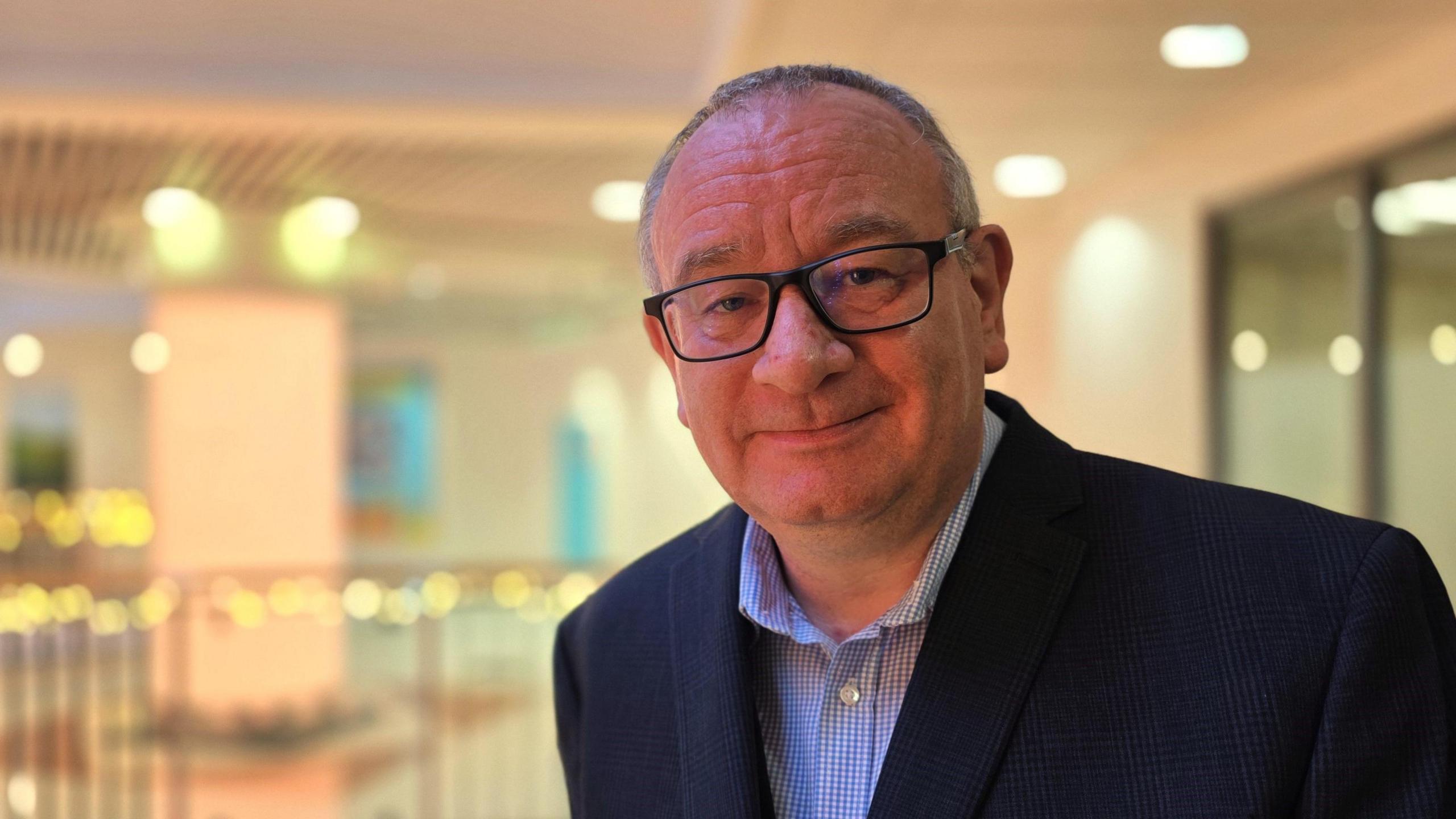 Rob Bradley is wearing a checked blue and white shirt beneath a navy suit jacket. He's also wearing glasses. Behind him we can a pillar, railings and various lights in the shopping centre he manages.
