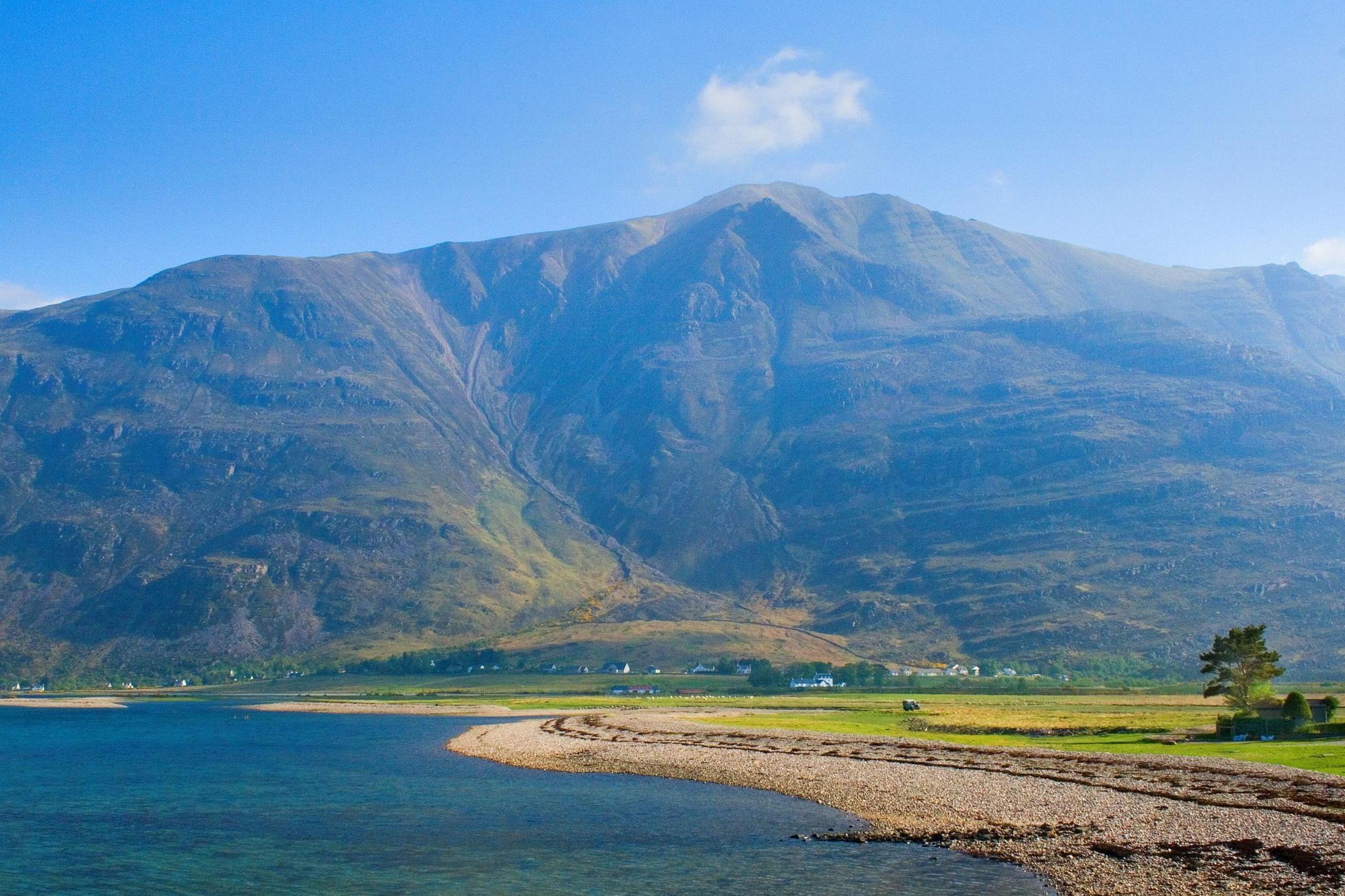 A view of Torridon on a sunny day. The small community is a scattering of houses along the shores of a sea loch and with large mountains rising behind it.