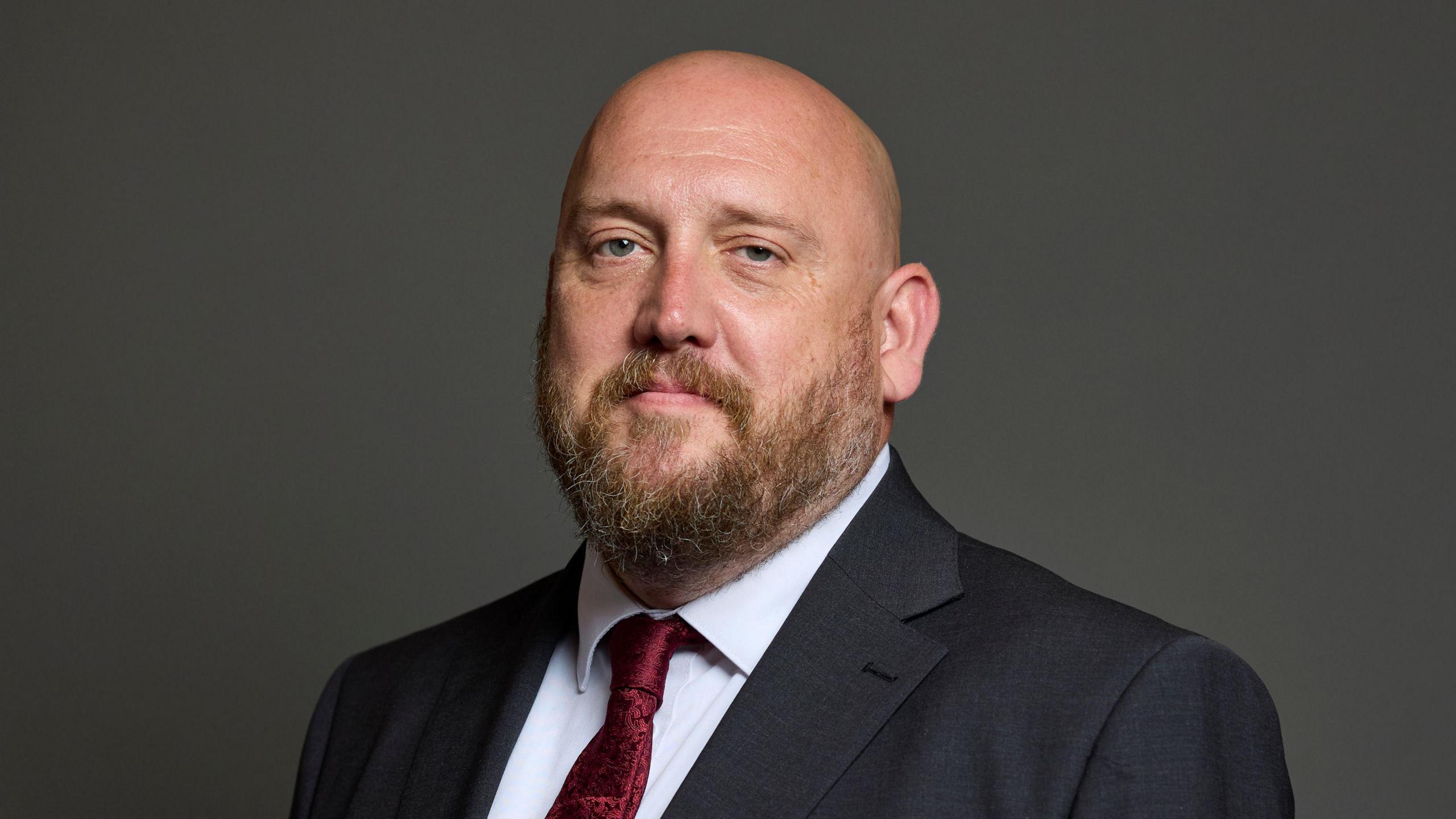 A bald man with a beard wearing a black suit and red tie.