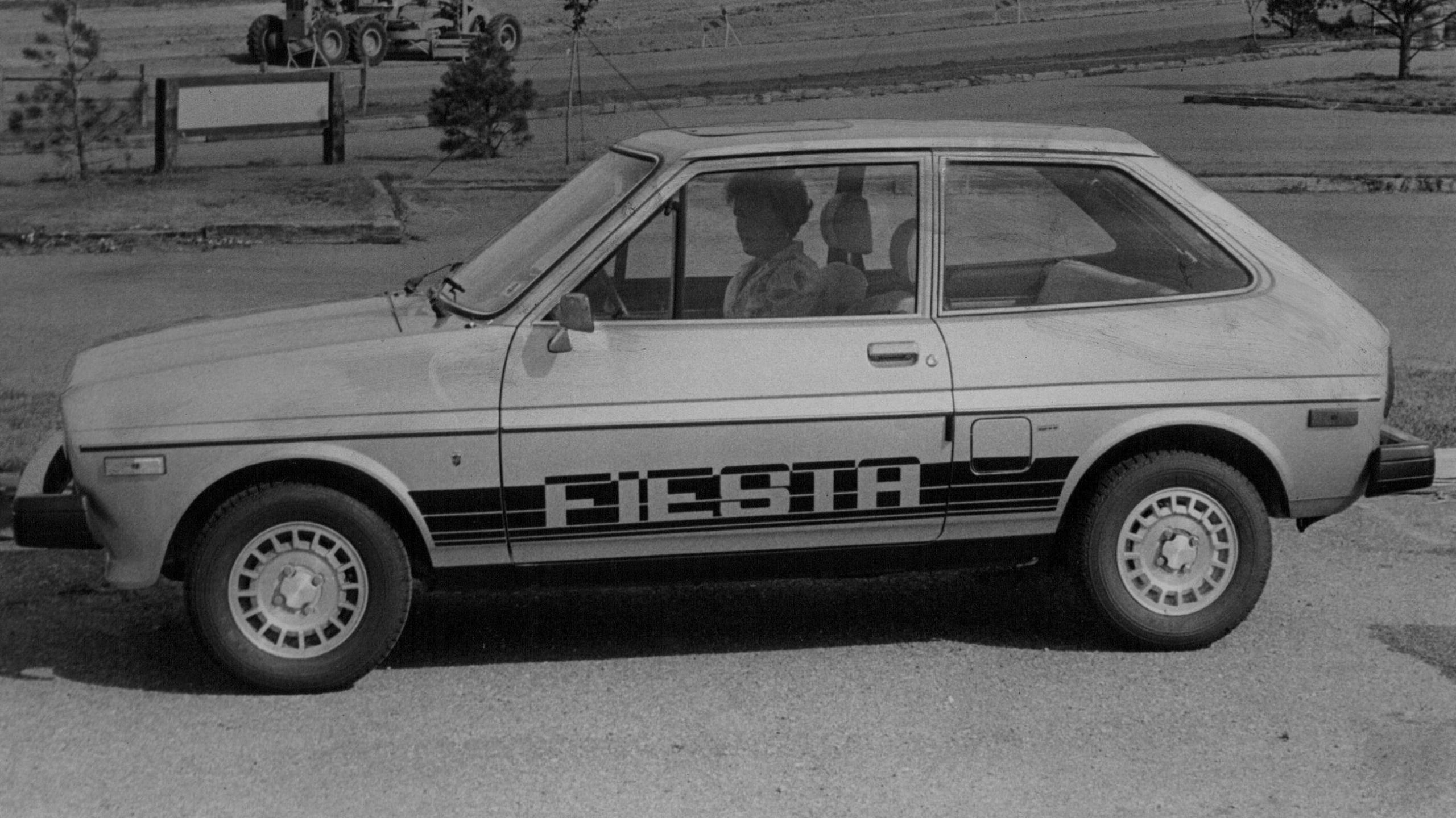 A black and white image of a vintage Ford Fiesta car. It has the words Fiesta written on the passenger door of the car. A woman is sat in the driver seat.