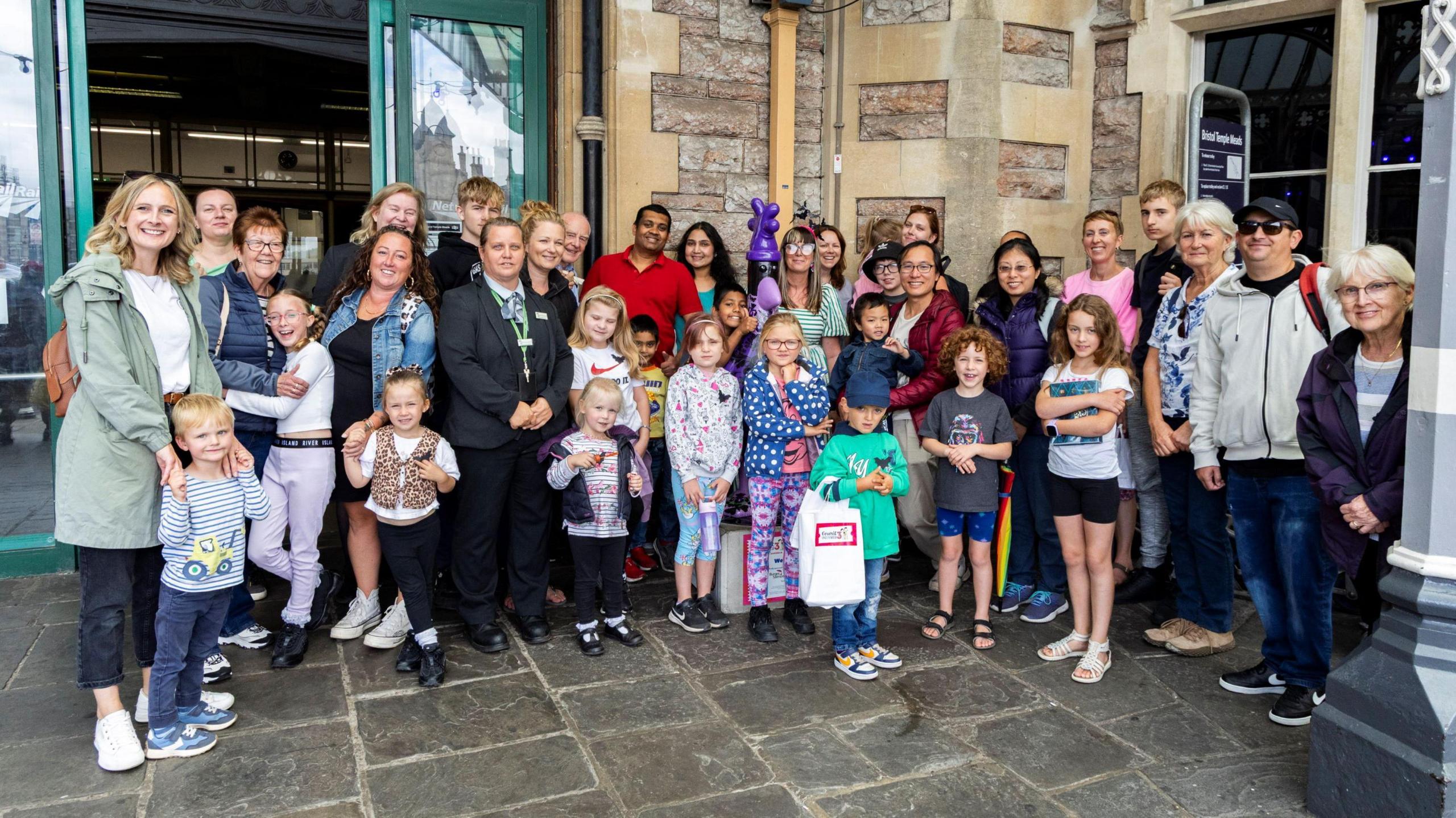 A large group of adults and children stand posing for the camera at the main entrance of Bristol Temple Meads train station. They are dressed for a rainy day.