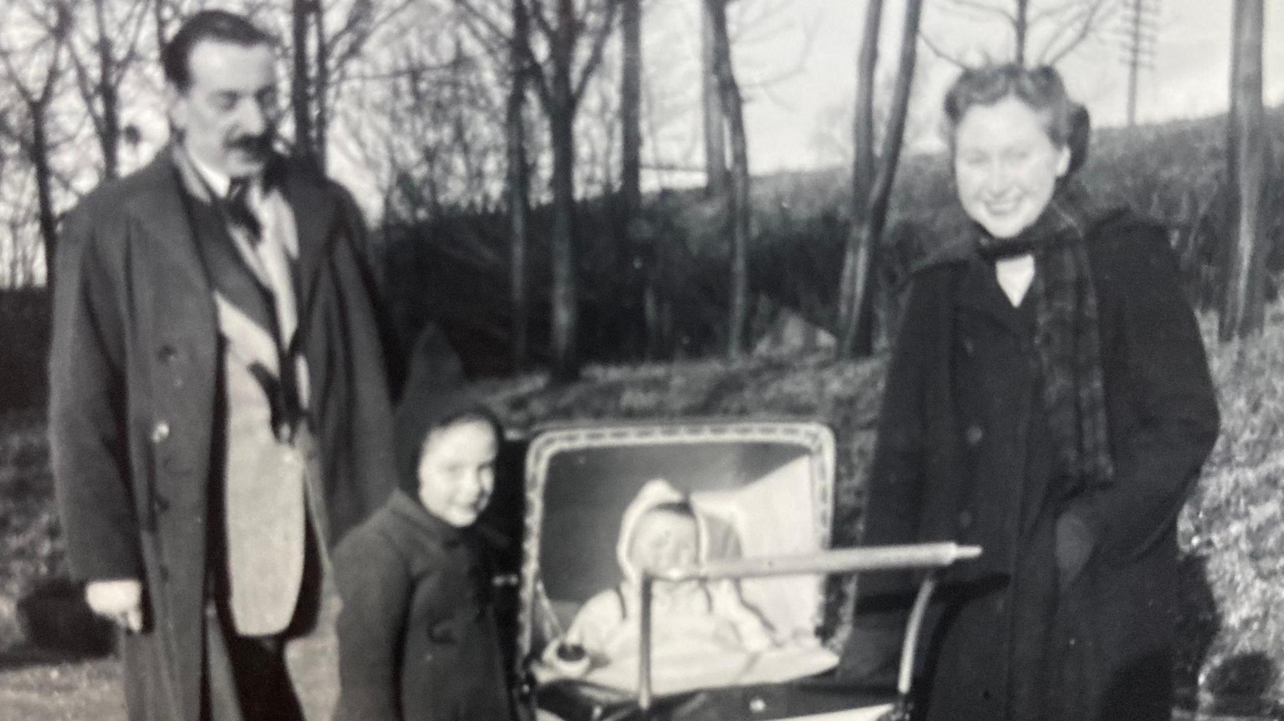 A black and white photo from the 1950s of Jan Hall's parents in a garden with a wooded area with a pram between them containing a baby and a little girl aged about 5 next to it.