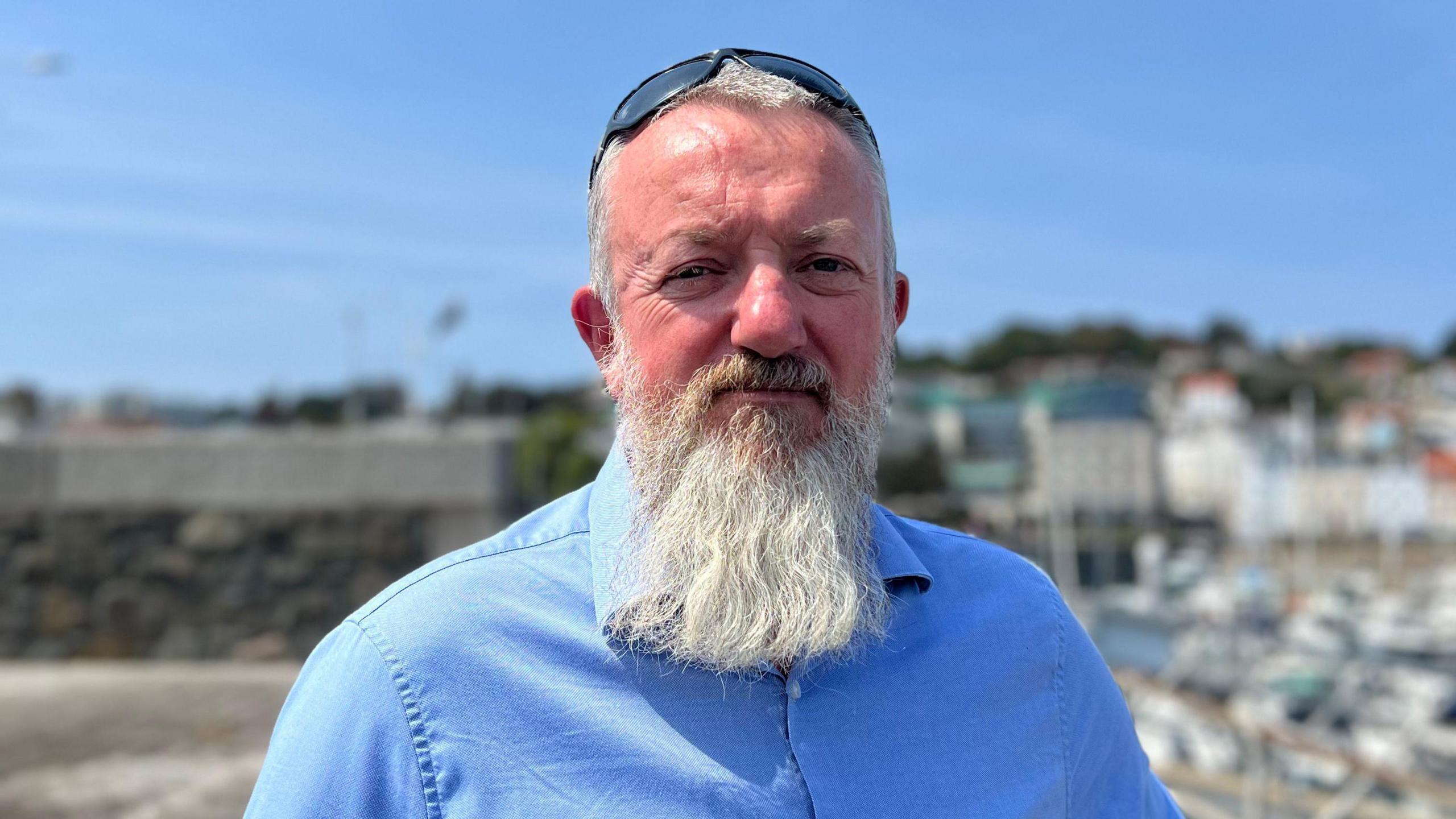 A man with a long gray beard and sunglasses on his head looks at the camera, wearning a blue shirt. 