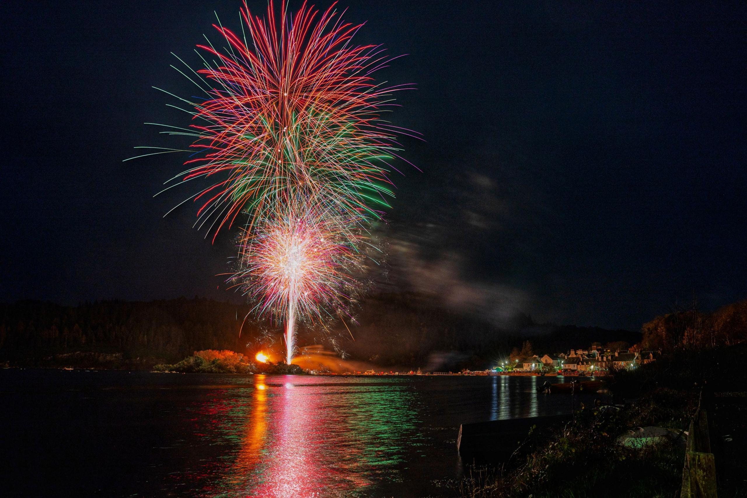 Brilliant fireworks explode in the night sky, with vibrant red and green streaks radiating outward in a dazzling display. Below, a quiet coastal village glows with warm lights, reflecting softly on the water’s surface.