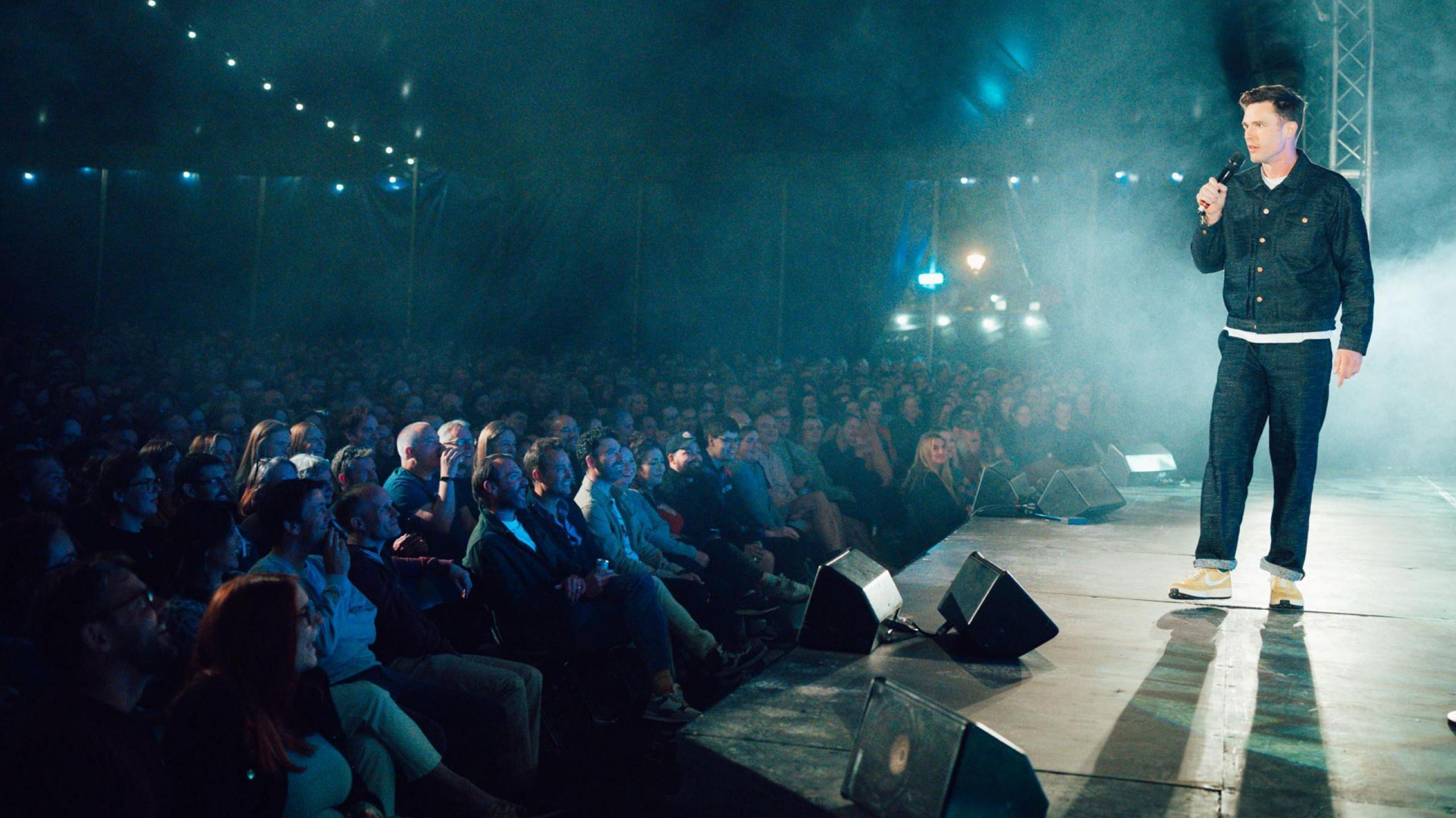 Comedian Ed Gamble, wearing dark jeans, a white T-shirt and a dark shirt, performs on stage at the Bristol Comedy Garden in Queen Square.