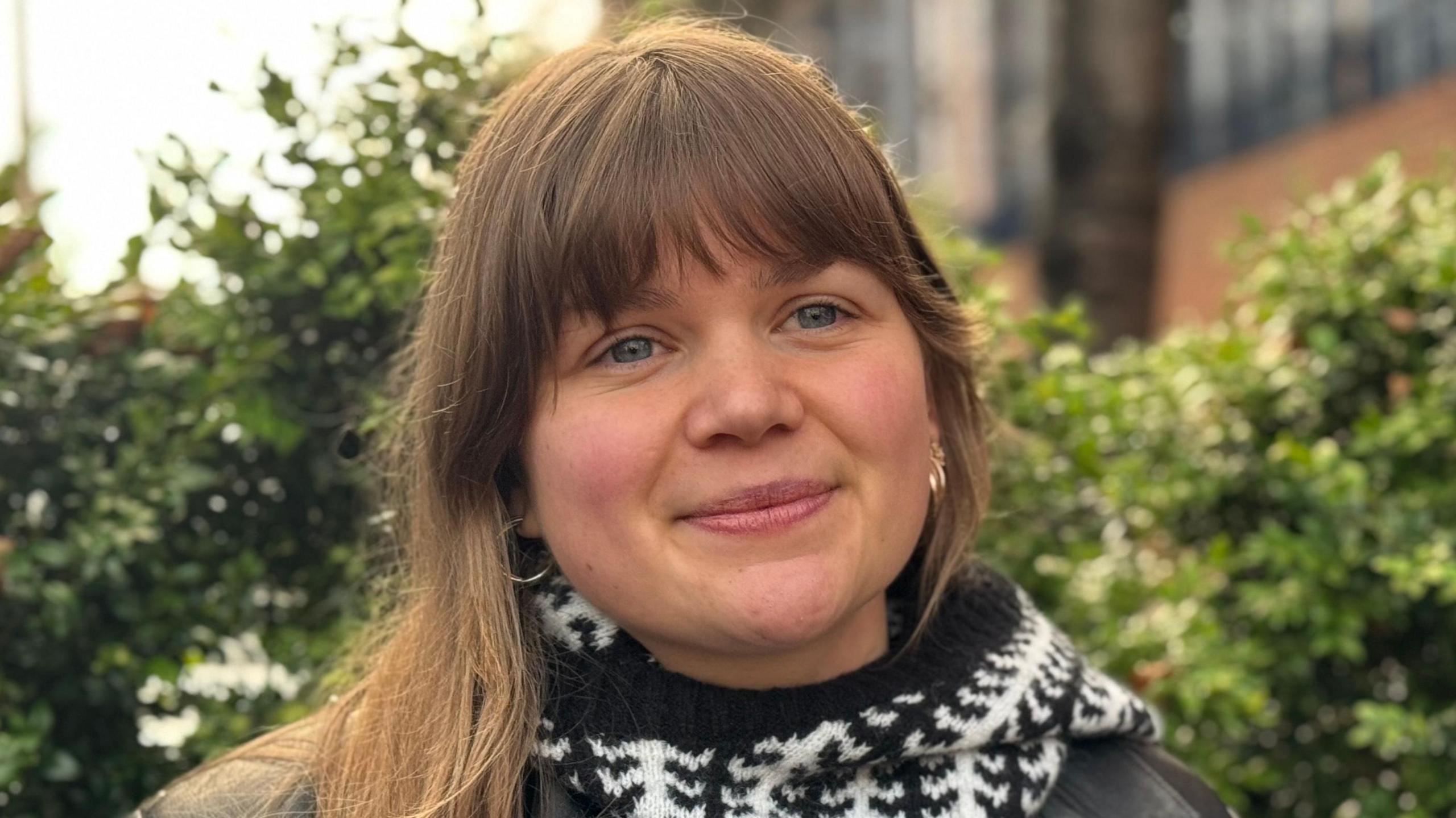 Polly Garnett looks to the camera. She has brown hair, with a straight fringe and is wearing earrings. She is wearing a black and white patterned scarf.