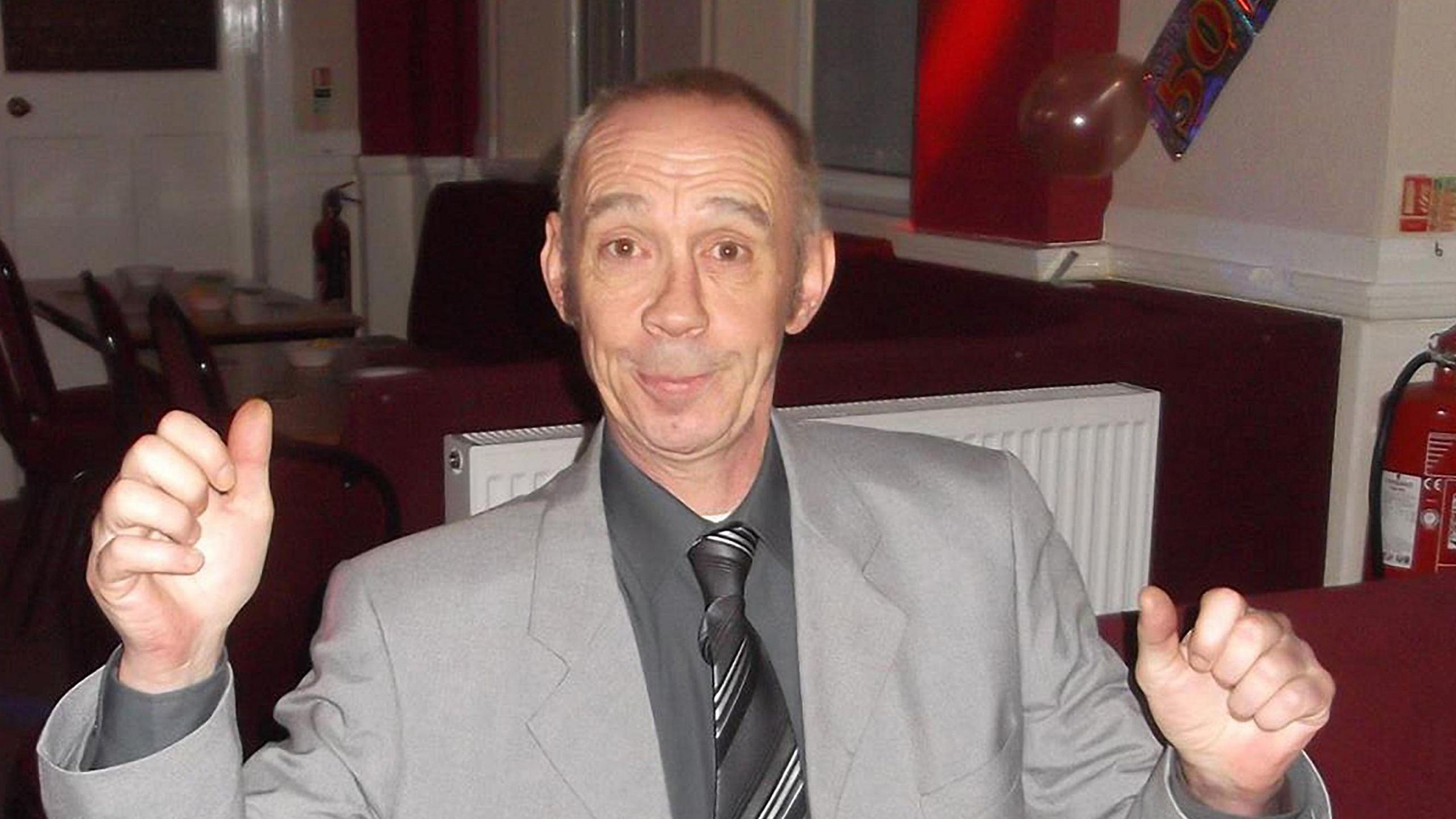 Ean Coutts looking at the camera. He has balding grey hair and is wearing a grey suit, grey shirt and grey and white striped tie. He is sitting in a social club/pub and is lifting his arms in the air.