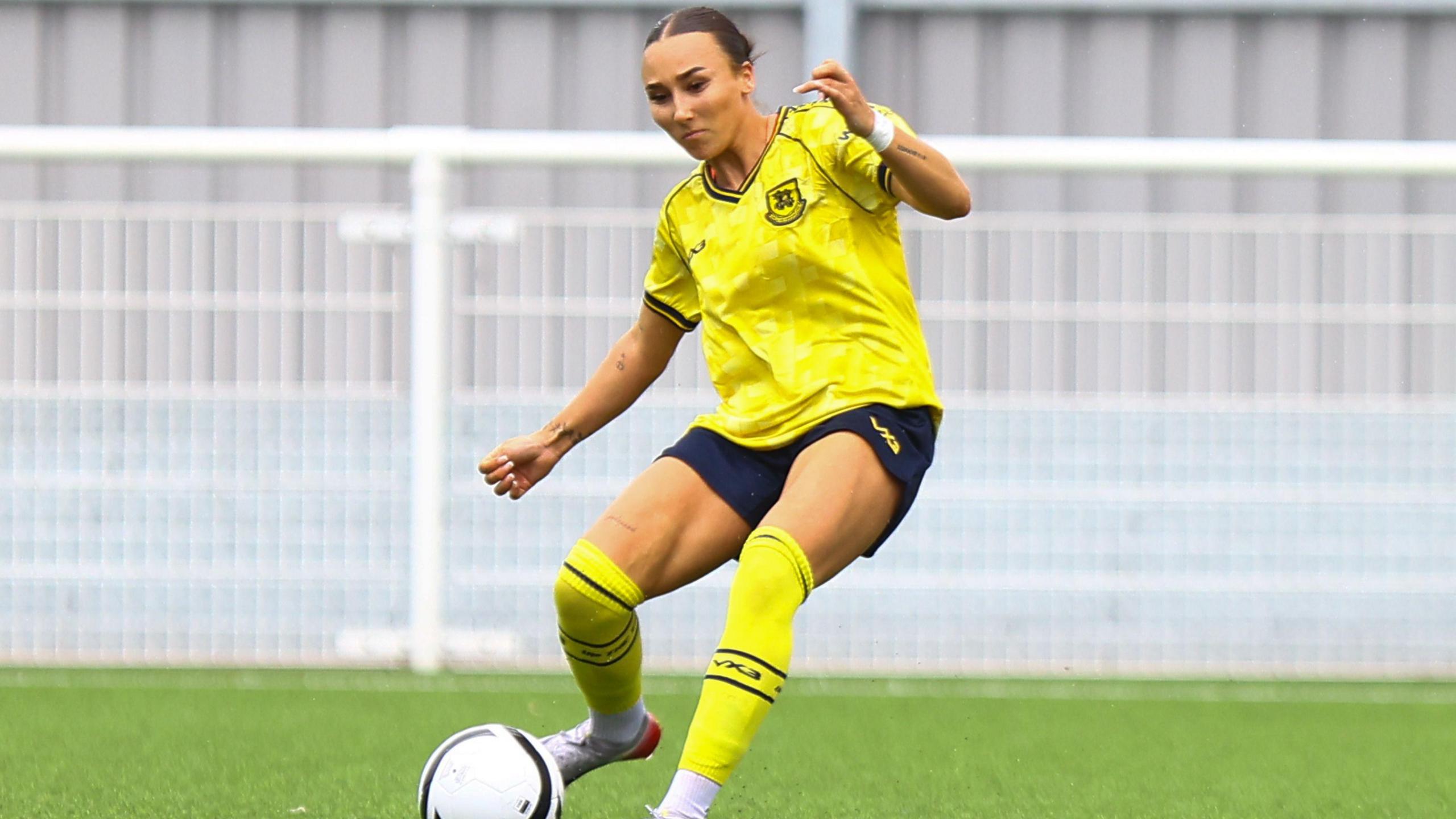 Connolly-Brame is wearing the yellow and blue Hashtag United kit, as she goes to strike a white football with her right foot. It is during a game..