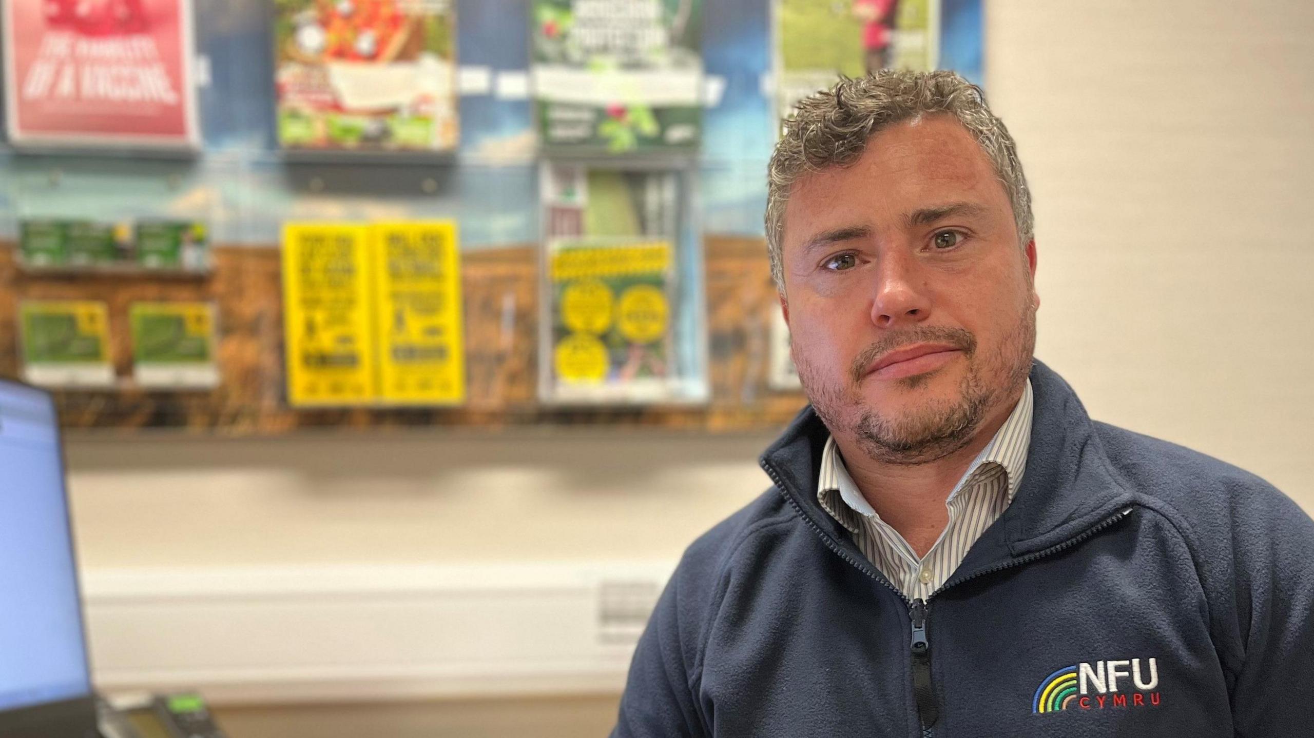 Aled Davies wearing a NFU Cymru-branded blue fleece, looks at the camera while sitting at a desk in an office