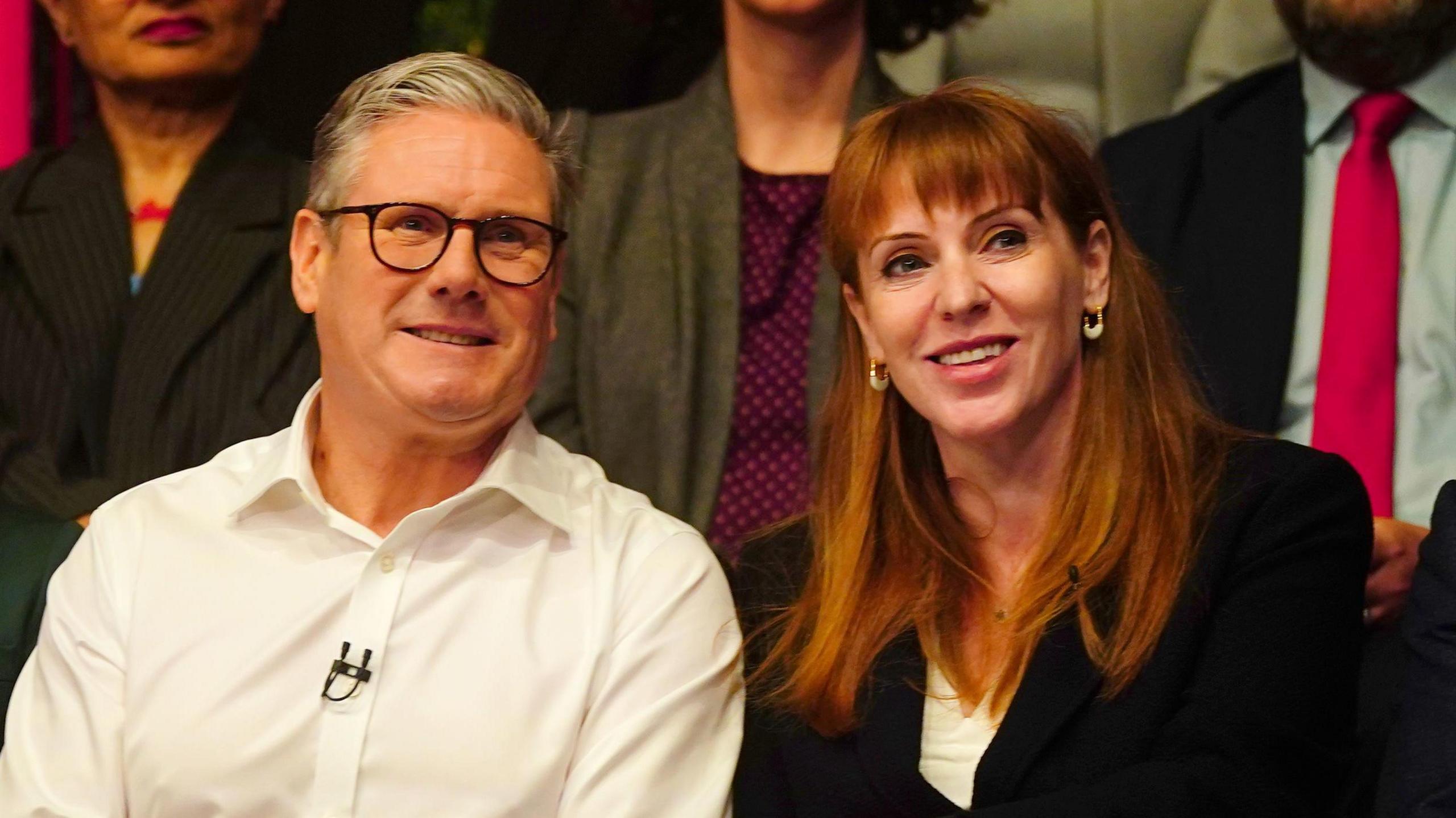 Starmer on the left in a white shirt with a mic pinned to it and Rayner next to him in a black blazer and white shirt. Both are sitting in an audience and smiling.