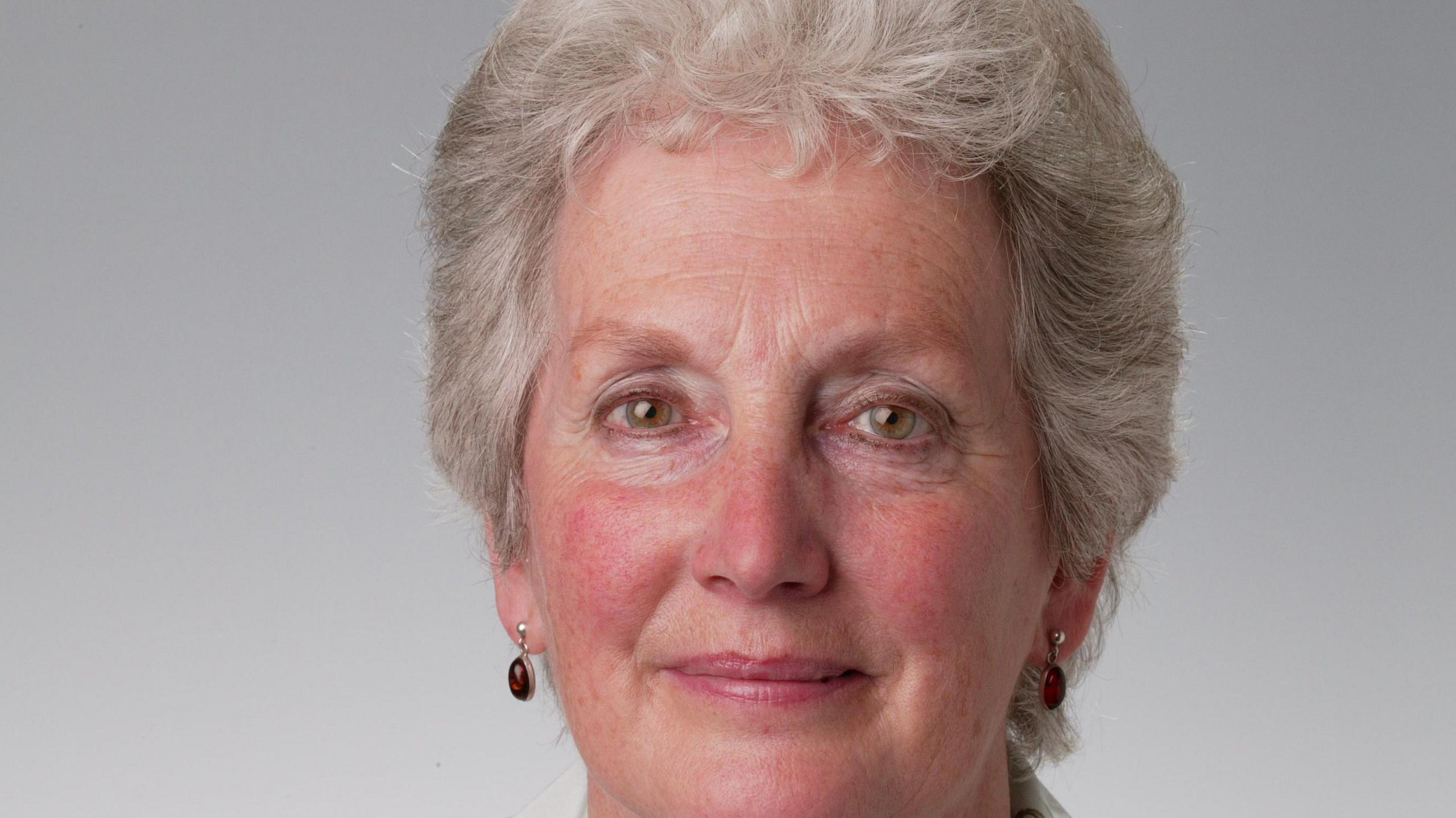 A picture of a smiling woman with short white hair. She is wearing gem earrings