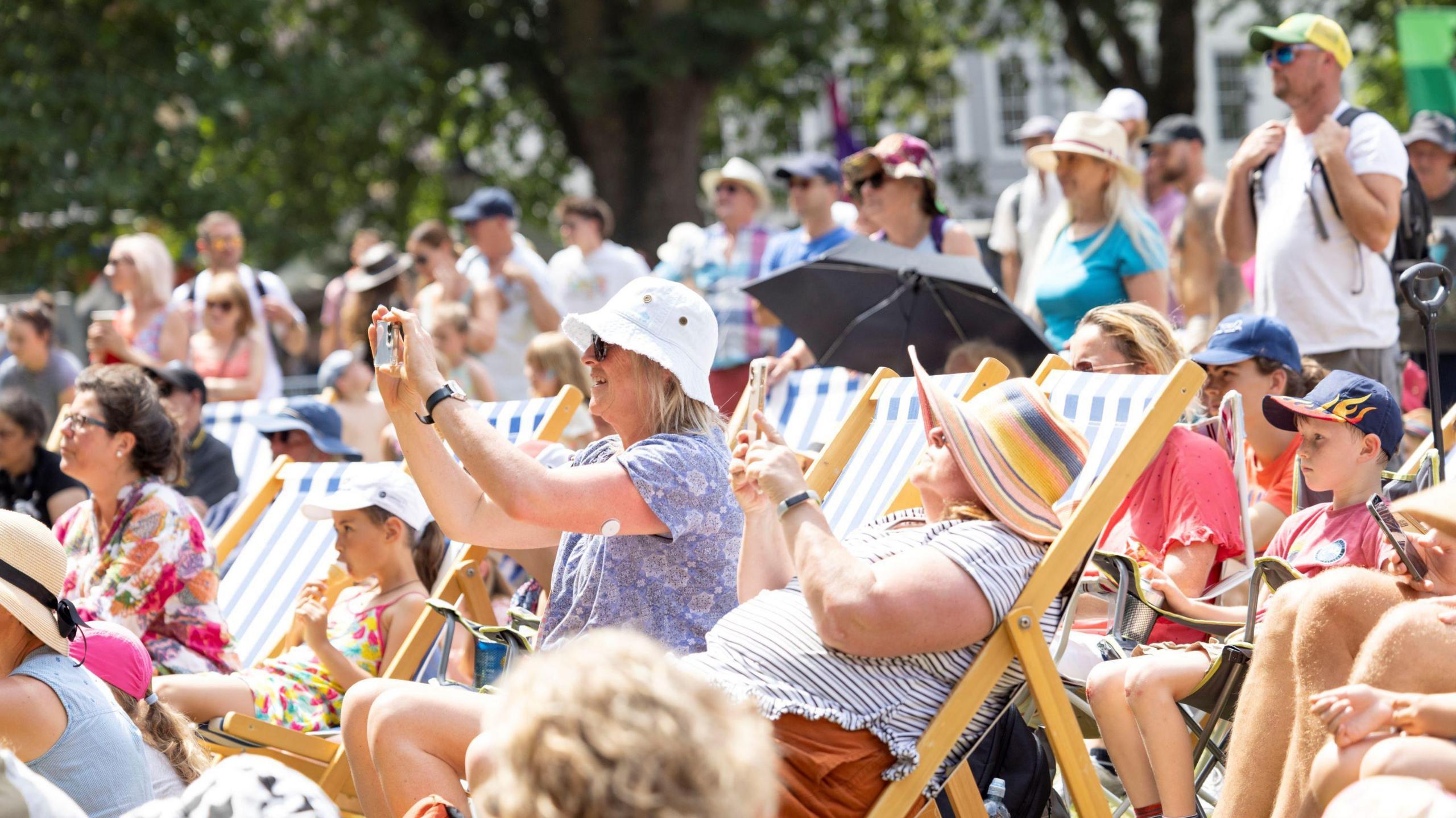 People sitting on deck chairs in the sunshine - a woman in the middle is taking a photo