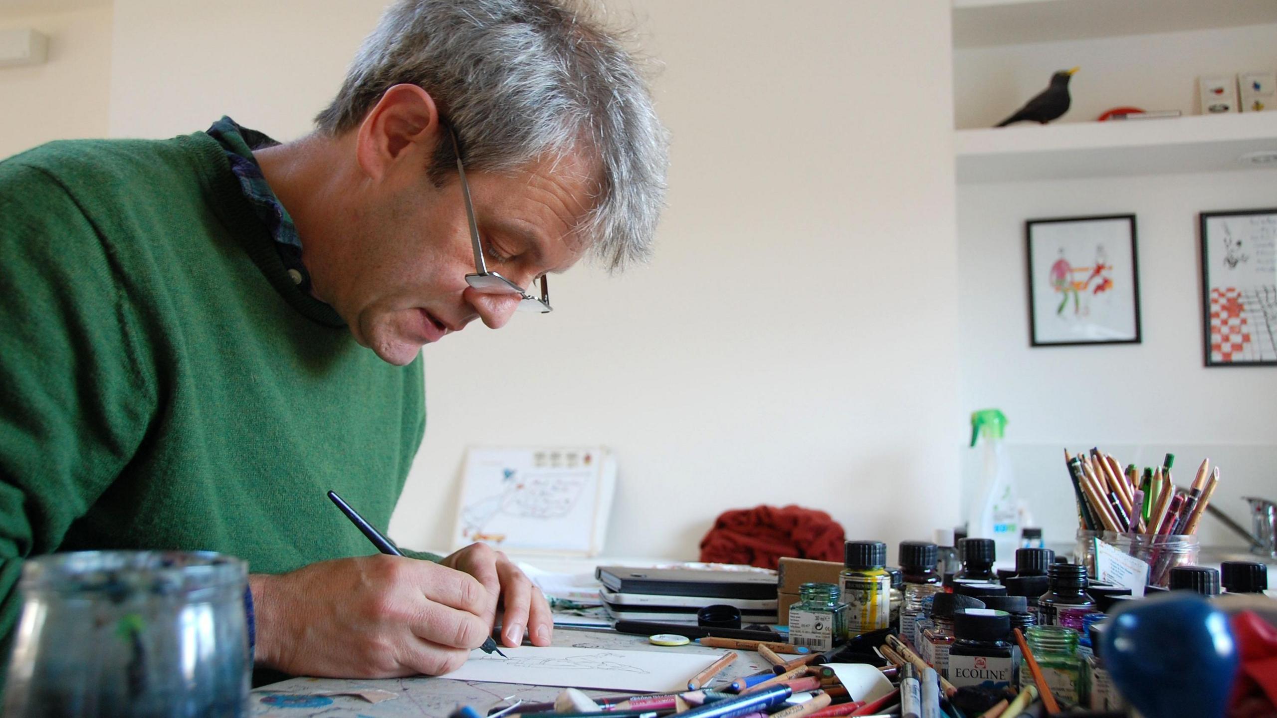 A man in a green jumper draws a picture on a desk full of paints and pencils