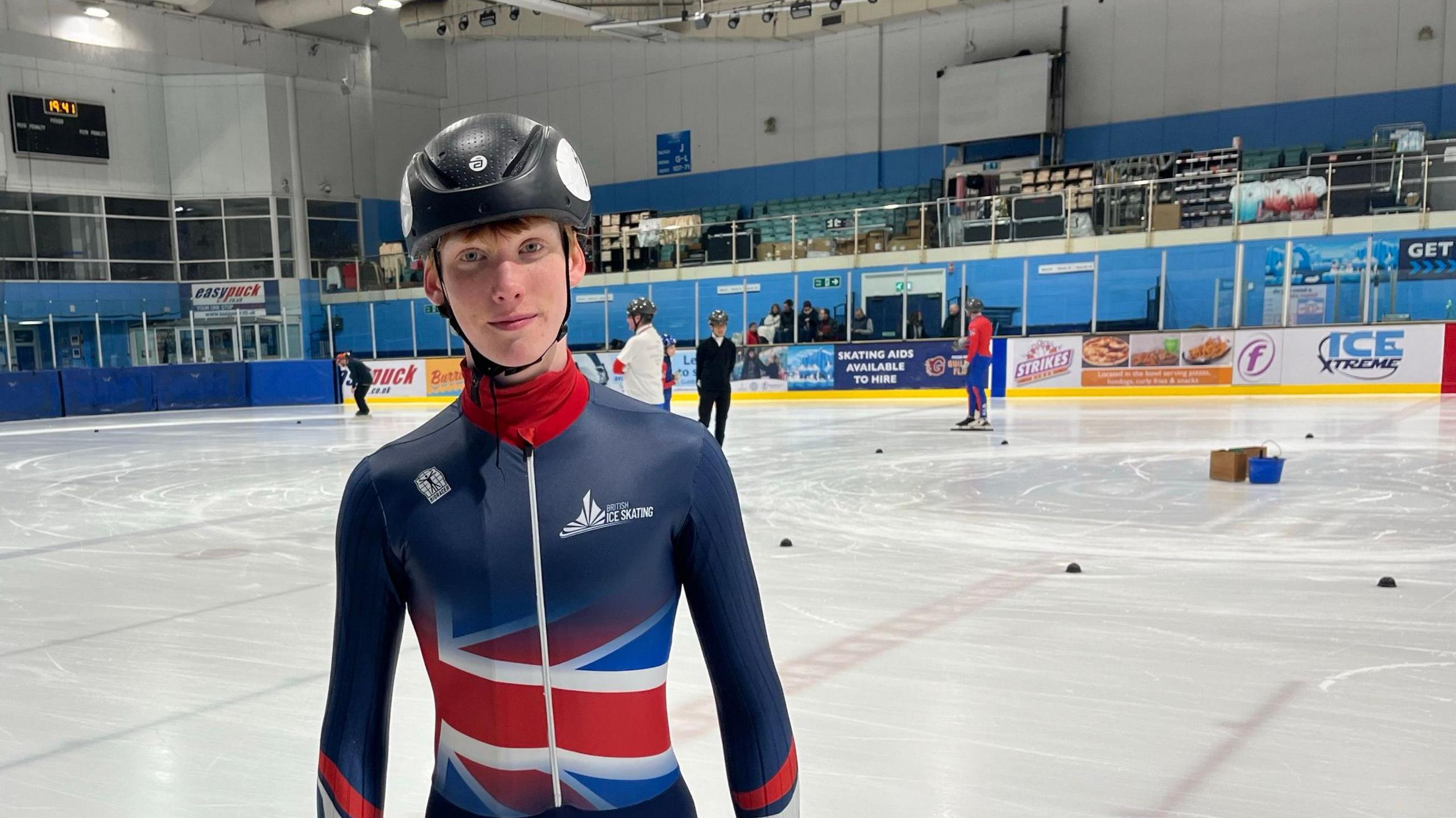 Albie Barlow, a teenage speed skater. He is stood on an ice rink wearing a black helmet and a navy blue leotard with a Union Jack flag on the front.