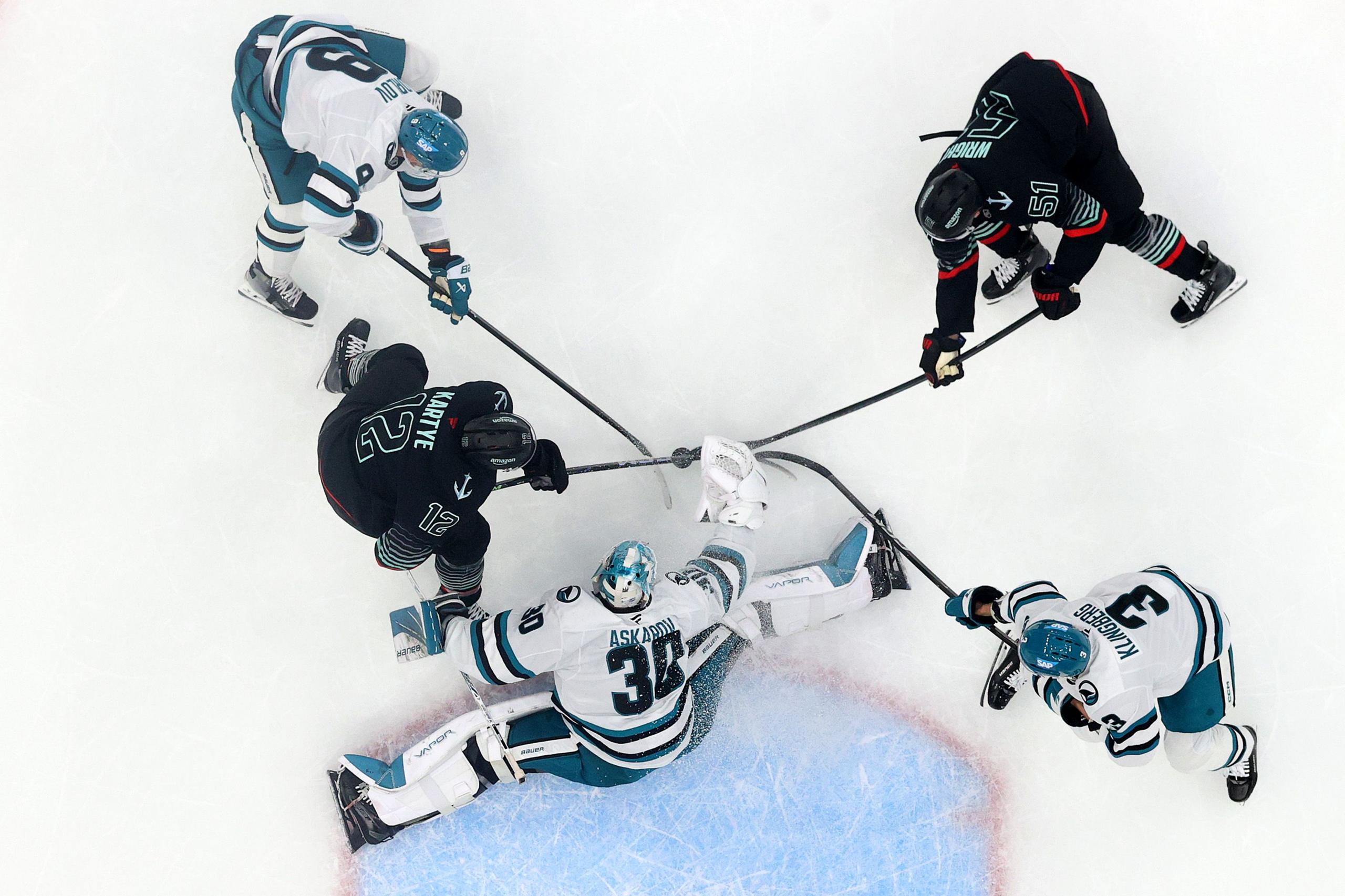 Top-down view of an ice hockey game showing five players and a goalie battling for the puck near the goal crease. Two players in black jerseys and three in teal-and-white jerseys are engaged with their sticks extended toward the puck, while the goalie in teal-and-white gear is sprawled across the ice making a save attempt.