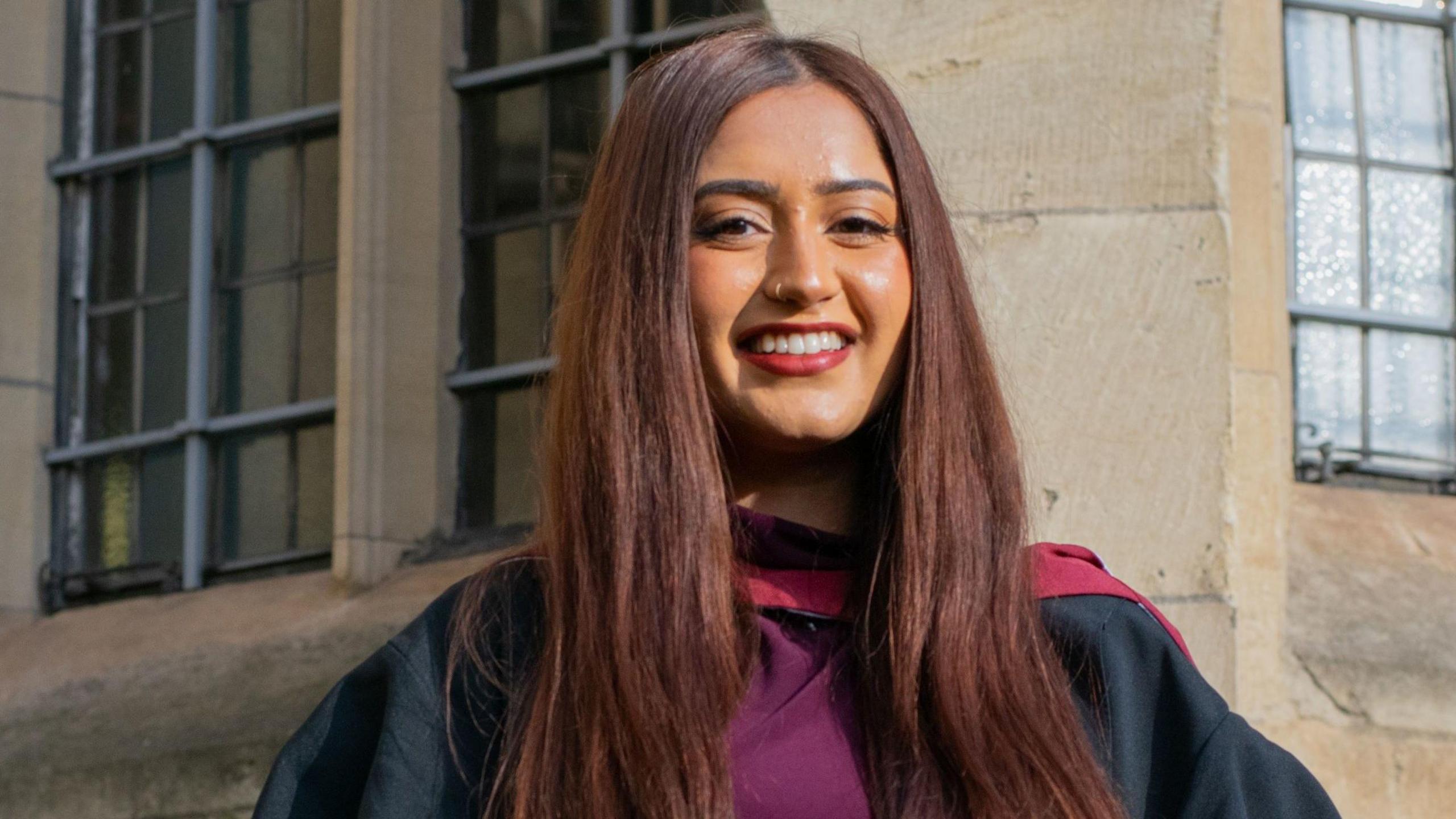 Bristol University graduate Gurvin Chopra smiles at the camera. She is wearing a purple and black graduation robe and has long dark brown hair. She is standing against a building with pale brickwork.