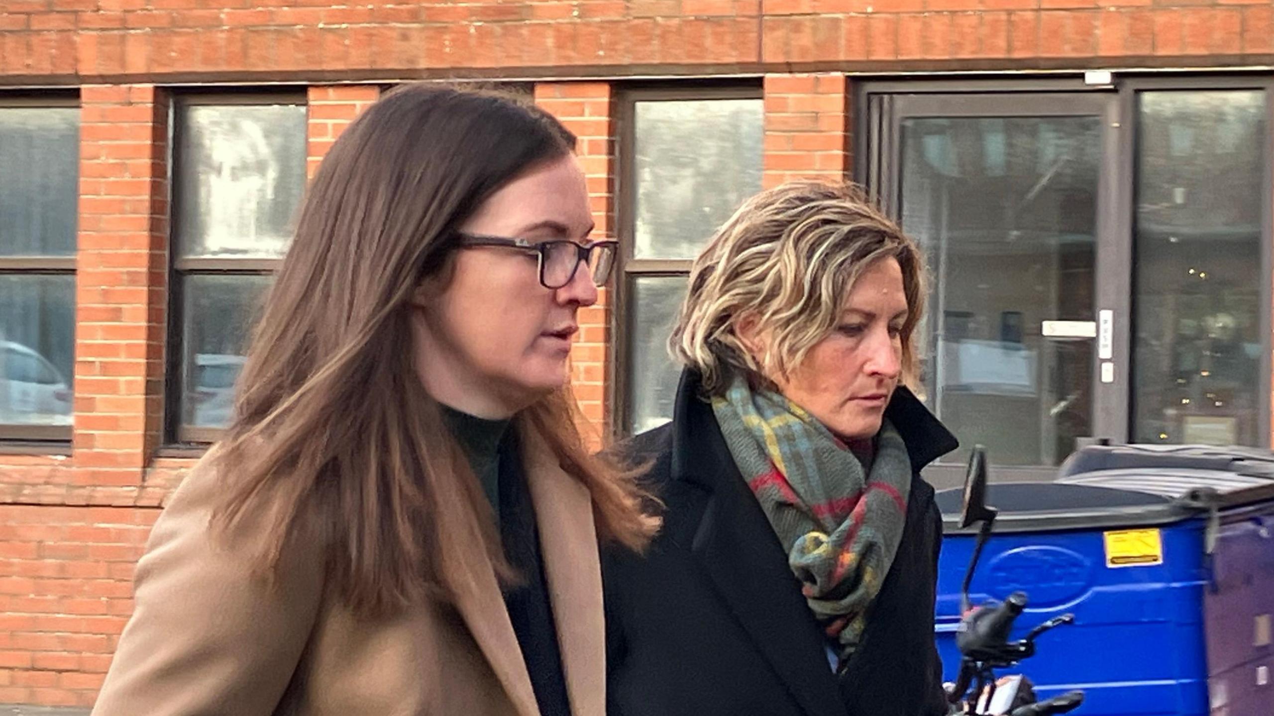 Two women walking alongside eachother past a red brick building. One is wearing a camel coat, glasses and has dark brown hair. The other, Nerys Lloyd, is wearing a dark coat and a multicoloured scarf. 